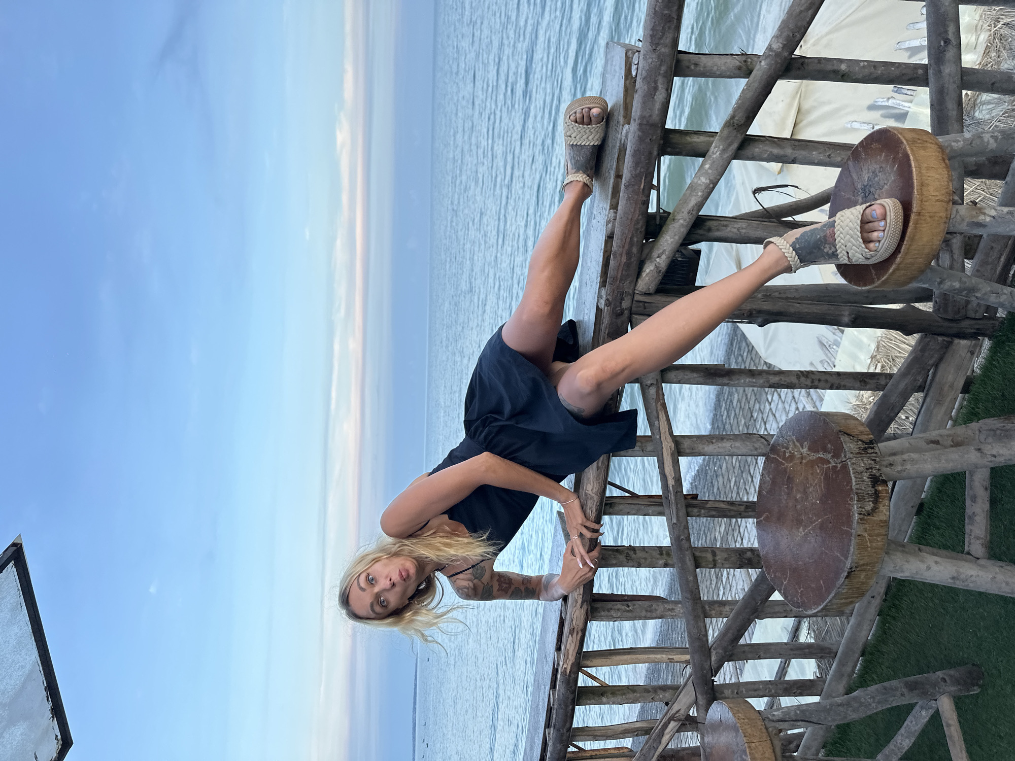A blonde woman is posing on a wooden railing overlooking the ocean. She is wearing a black dress and sandals. The woman's left arm is resting on the railing, and her legs are extended. Her facial expression is playful. In the background, the ocean is calm, and the sky is a pale blue with wispy clouds. There are also wooden stools visible in the foreground. The image appears to be taken at dusk. The location is identified as Phu Thuy Ward, Vietnam, suggesting a coastal resort or dining area.