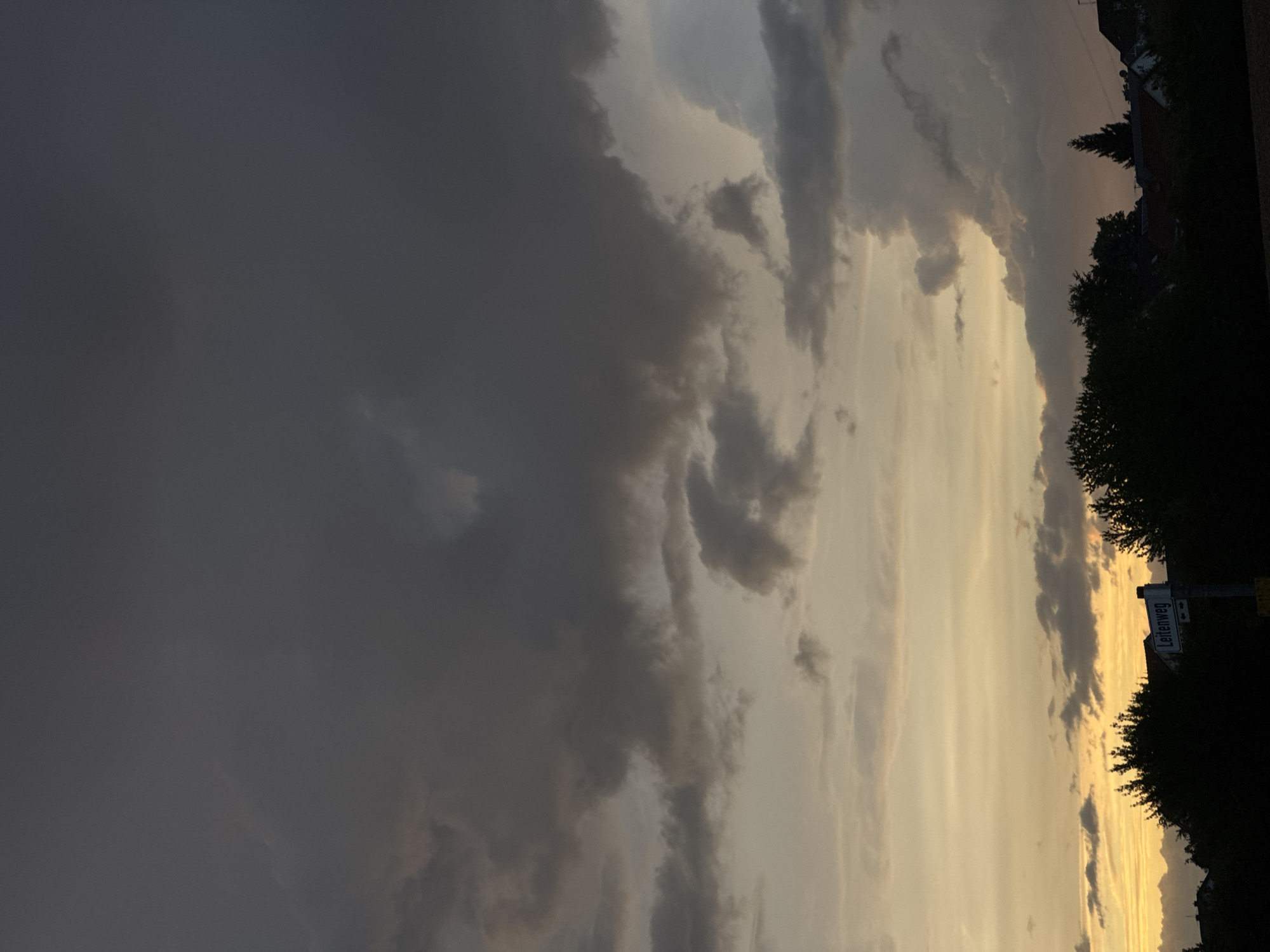 The image depicts a dramatic sky over Wettstetten, Germany, during what appears to be sunset or a storm approaching. Dark, heavy clouds dominate the upper portion of the frame, with lighter, more diffused clouds visible lower down, tinged with yellow and orange hues from the setting sun. Silhouetted against this sky are trees and the rooftops of houses, indicating a semi-rural or suburban setting within the town. A street sign that reads "Leitenweg" is visible on a post in the foreground, further confirming the location. There are no people or active events captured in the photograph; it is a static scene focusing on the natural elements and the built environment. The overall mood is one of impending weather or the transition from day to night.