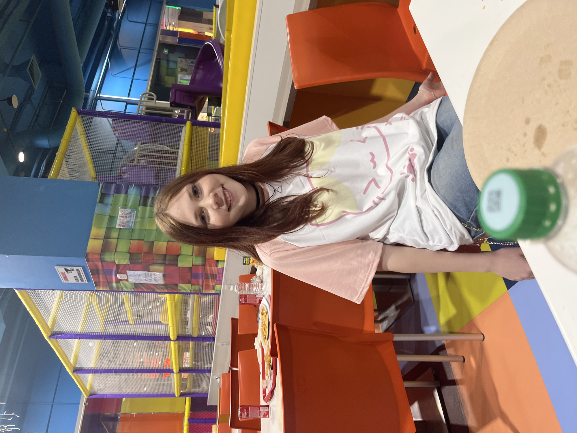 A young girl sits at a table with orange chairs, likely in a play area or children's center in Sochi, Russia. The background features colorful play structures with netting and padded elements. The girl wears a light pink and white T-shirt and jeans. A pizza and bottle with a green cap are visible on the table. Other tables and chairs extend behind her.