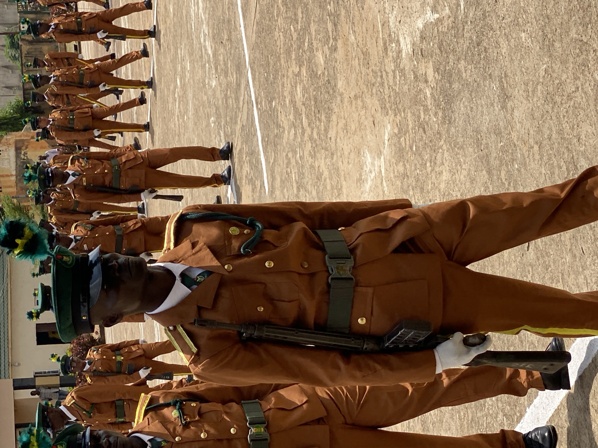 This image depicts a military parade or training exercise taking place outdoors on a paved surface, likely a parade ground, in Kaduna, Nigeria. A group of individuals, all male and appearing to be soldiers or cadets, are lined up in formation. They are wearing matching brown uniforms with green accents on their hats, belts, and lapels. Many of them are holding rifles, suggesting a drill or ceremonial event. The time of day appears to be daytime, with bright sunlight casting shadows, indicating fair weather. The background shows some buildings and greenery, consistent with an outdoor location in Nigeria. The soldiers are mostly facing forward or to the side, with a somber or focused demeanor, typical of such events. One soldier in the foreground is clearly visible, wearing a green cap with a feather plume. The overall scene conveys discipline and order. No visible text is present in the image.