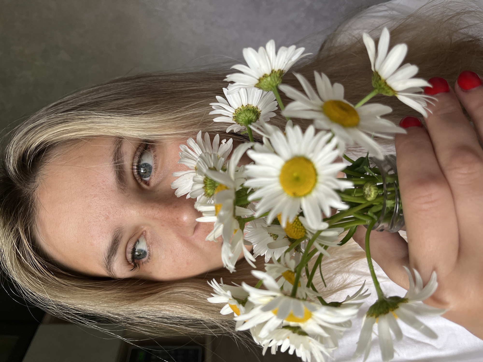 A close-up, eye-level, medium shot shows a young blonde woman holding a bouquet of daisies. Her face is partially obscured by the flowers, but her blue eyes are visible as she looks to her left. She has light skin with freckles and her hair is a light blonde color, falling around her face and shoulders. Her fingernails are painted a bright red. The daisies are white with yellow centers. The background is a textured gray wall. The lighting is natural and appears to be coming from the left. The scene is indoors. There is no discernible location in Chaikovsky, Russia.