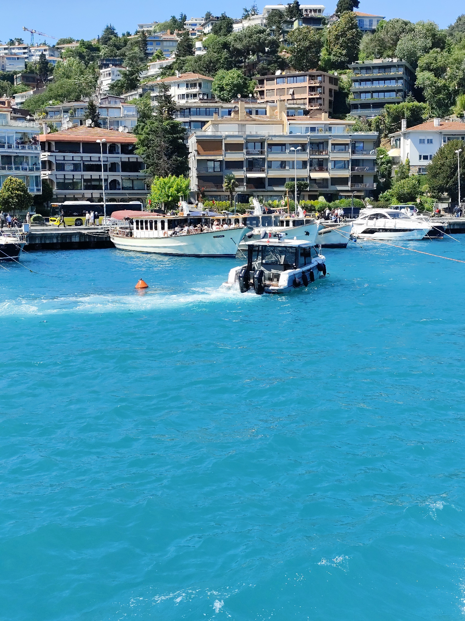 The image depicts a waterfront scene in Beşiktaş, Turkey, characterized by bright blue water filling the majority of the frame. In the background, a steep hillside is densely populated with multi-story buildings and trees.
Several boats are docked along the pier, with passengers visible on the decks. A bus is visible behind the docked boat, possibly for transporting tourists. An orange buoy floats in the water near the docks.
In the foreground, a small, motorized boat is in motion, leaving a trail of white wake in the blue water. The stern of the boat, with visible twin outboard motors, faces the viewer. The overall impression is of a busy, tourist-oriented area on a sunny day.