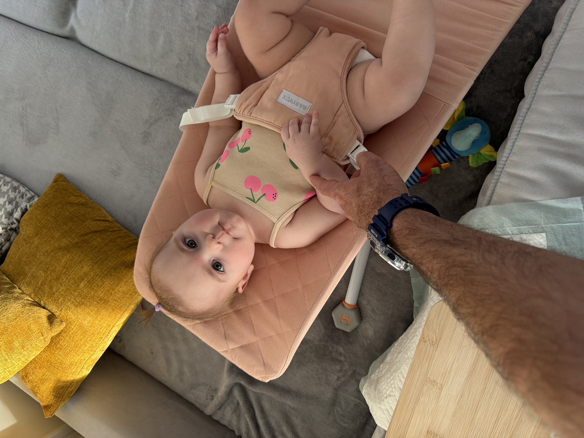 An overhead indoor shot captures an infant lying in a peach-colored baby bouncer, with an adult's arm extended towards the baby's right arm. The setting appears to be a comfortable home environment, possibly a living room or nursery in Krasnoyarsk, Russia, suggested by the surrounding furniture.
The baby, with fair skin and light hair styled in two small pigtails, looks directly at the camera with a calm, observant expression. They are wearing a light-colored, possibly beige, onesie adorned with pink cherry or strawberry motifs. A beige harness from the bouncer secures the baby, featuring a label with the text "BABYROX". The bouncer itself has a textured, quilted fabric.
To the left of the bouncer, a grey sofa is visible, partially covered by a white and grey patterned blanket and adorned with two yellow throw pillows. To the right, part of a wooden tray or board is visible, along with white and light green fabric, possibly linens or burp cloths. Underneath the bouncer, on a grey carpeted or upholstered floor, a colorful blue elephant-like toy is partially visible.
The adult's arm, hairy and wearing a blue digital watch, gently touches the baby's arm. The watch face shows the text "G-LIDE" and "30M". Part of the bouncer's base, a grey leg with an orange "H" symbol, is also visible near the adult's hand. The lighting suggests it is daytime. The overall scene depicts a gentle interaction between an adult and a baby in a relaxed home setting.