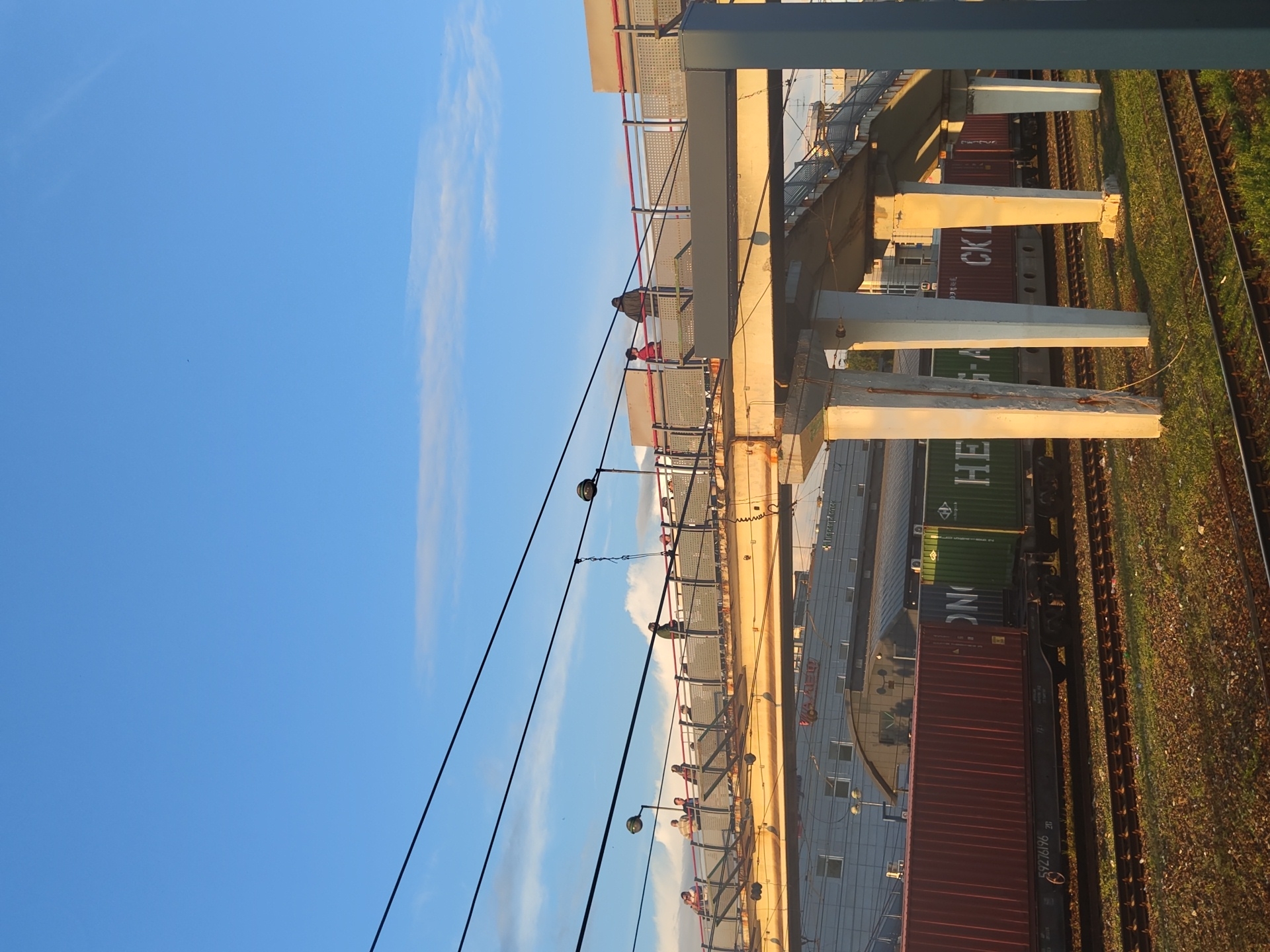 An eye-level, wide shot captures a pedestrian overpass spanning a railway track at sunset in Ramenskoye, Russia. The sky is a brilliant, cloud-streaked blue. Below the overpass, a train carries several red and green shipping containers. The overpass, constructed of concrete and metal, is worn and weathered. People are seen walking on the overpass, some looking down at the train. The scene is bathed in the warm, golden light of the setting sun, casting long shadows.