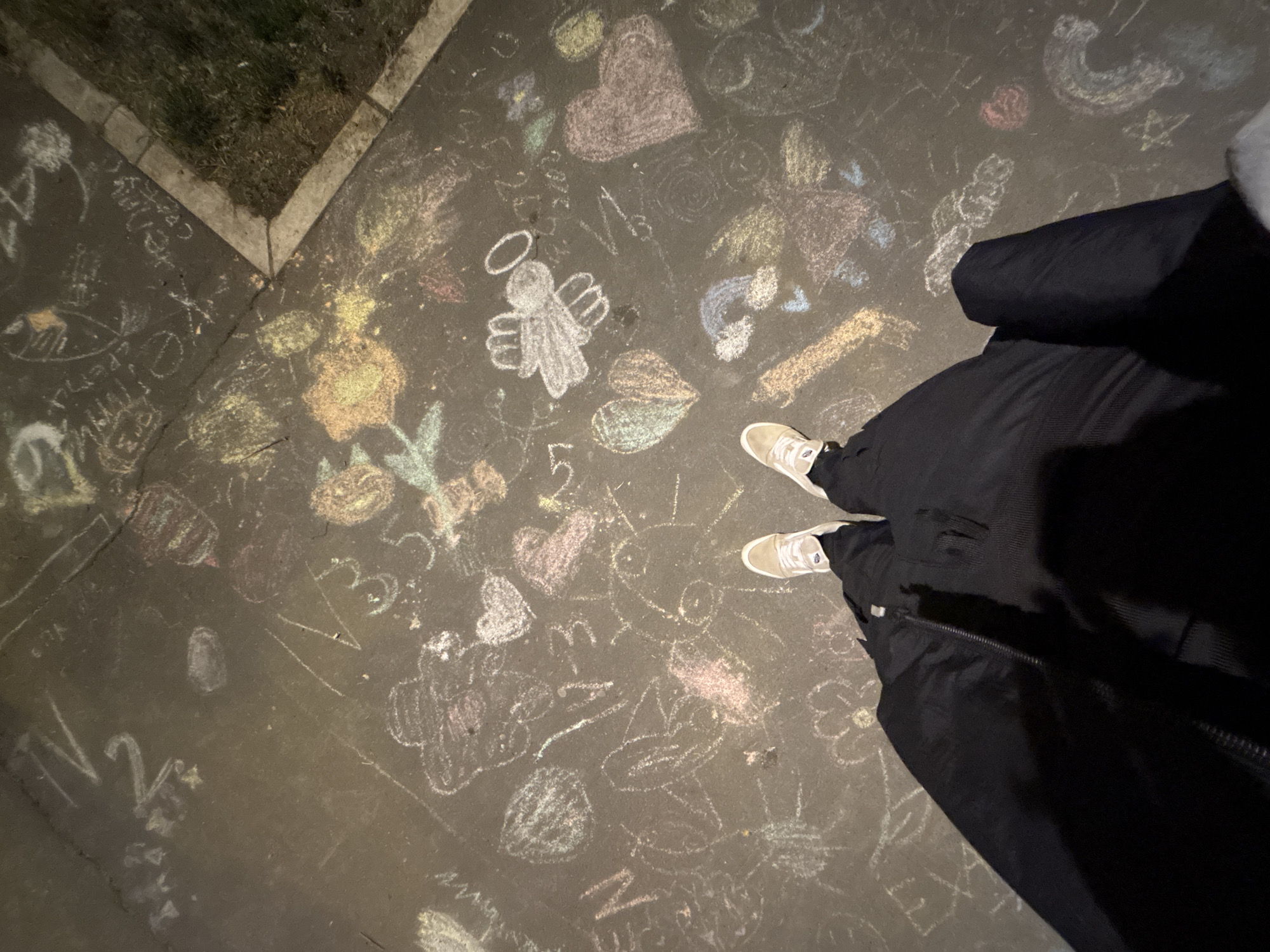 A photograph shows a section of pavement covered in chalk drawings. The drawings include hearts, flowers, numbers, letters, an angel, and other shapes. A person wearing light-colored sneakers and a black winter coat is standing on the pavement. The photograph appears to have been taken at night, judging from the light and shadows. A small patch of grass is visible at the edge of the pavement. The location is not discernible from the image alone and therefore Palilula Urban Municipality, Serbia cannot be confirmed.