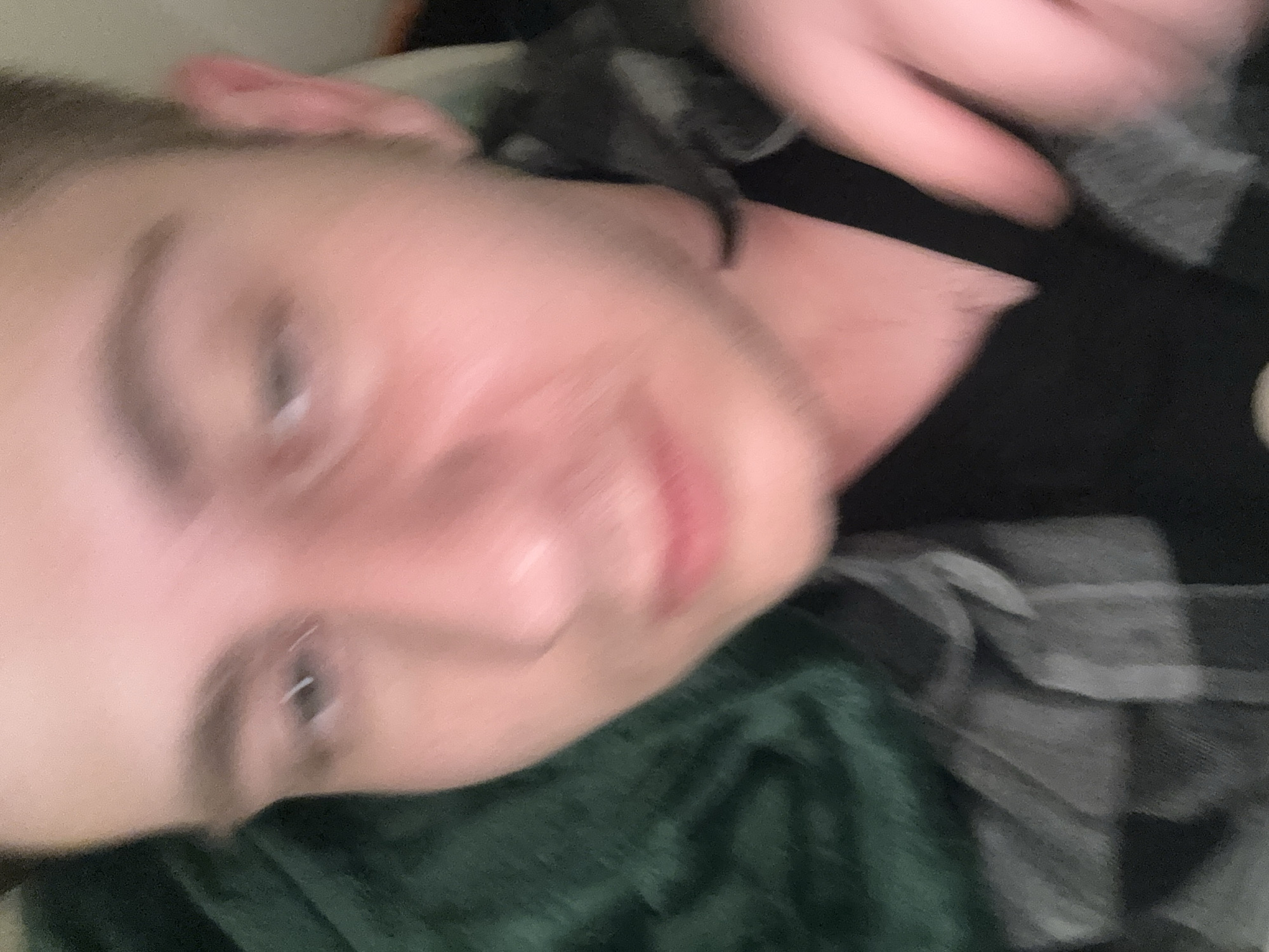 The image presents a blurry, close-up shot of an individual, likely a young adult, lying down indoors. The person has fair skin, short hair visible on the side of their head, and light-colored eyes. Their lips are slightly parted, and a hand is visible, blurry, in the upper right corner of the frame.
The individual is dressed in a black undershirt, worn beneath a plaid shirt with a grey and black checkered pattern. A dark green blanket or fabric is draped across the lower portion of their body. The overall setting appears to be an interior space, possibly a bedroom or living room, characterized by low light conditions and indistinct background elements due to the blur.
The activity suggests the person is resting or relaxing. The blur throughout the image implies either subject movement, camera shake, or a slow shutter speed in dim lighting. No specific emotions are clearly discernible due to the blur, but a relaxed state is suggested. There are no visible location cues, text, or explicit indicators of the time of day or weather within the image.