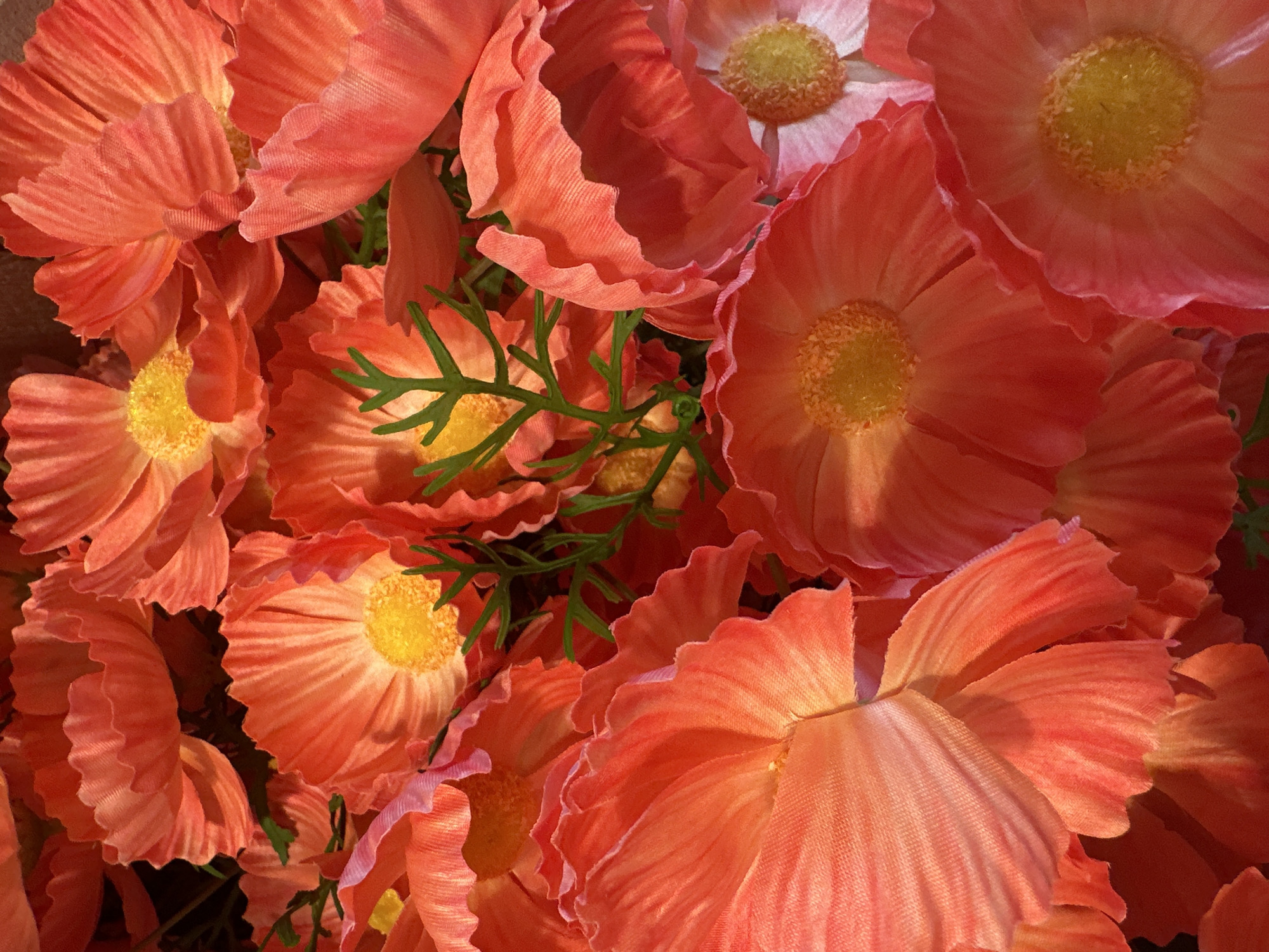 The image is a close-up, overhead shot of a cluster of artificial poppies, predominantly in shades of orange and pink. The flowers have delicate, ruffled petals and bright yellow centers. Some green stems and leaves are visible amongst the blooms, suggesting a floral arrangement or bouquet. The lighting creates highlights and shadows that emphasize the textured surfaces of the petals. There is no indication of a specific location in Steenwijk, Netherlands, or any people or actions depicted.