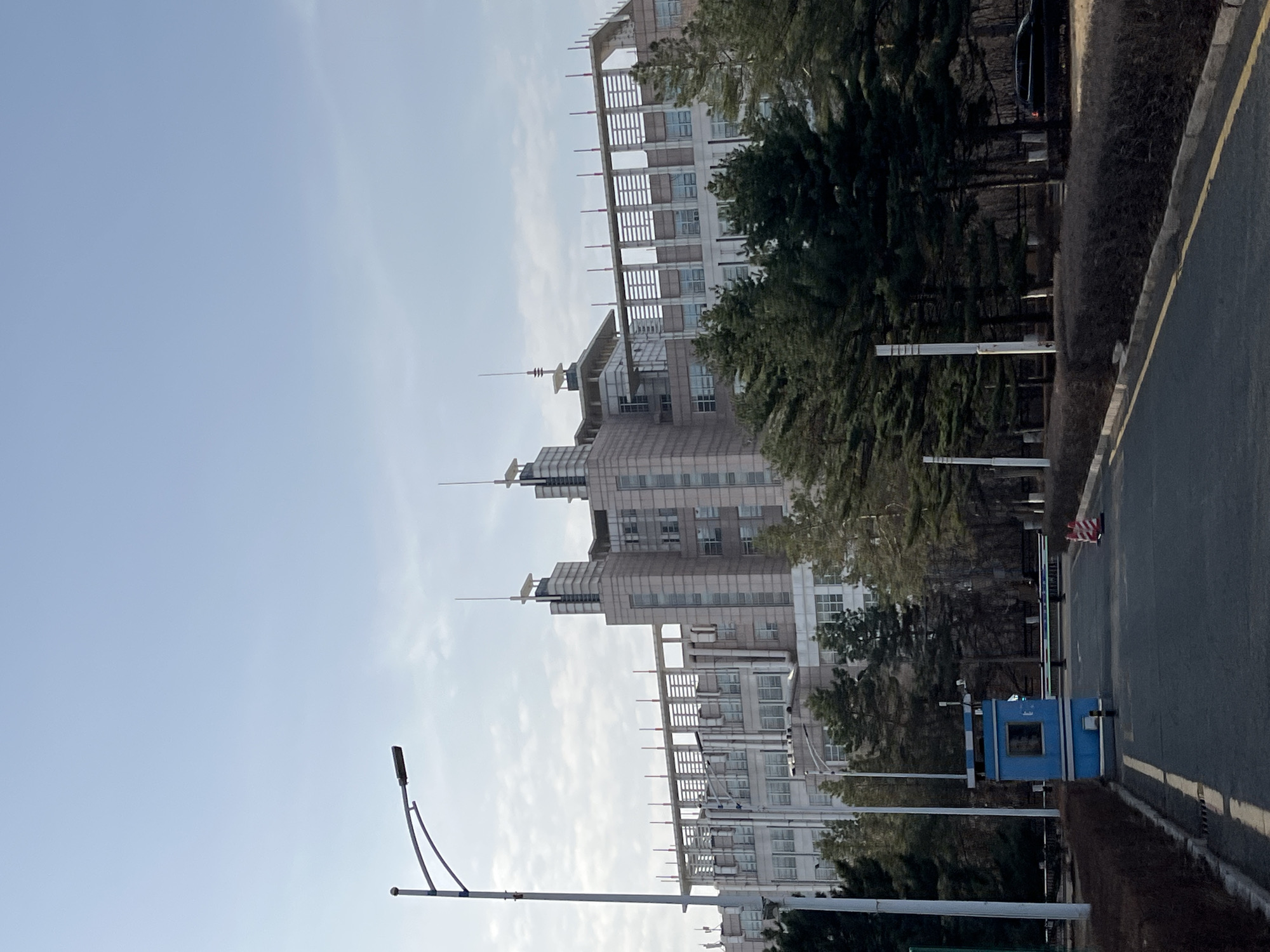 The image shows a view of a building in Changchun City, China. The building is tall and constructed with multiple stacked segments, with white windows, balconies, and antennae. Trees partially obscure the building from the roadside. A street runs along the right side of the image. There is a blue security booth and a traffic control arm. A lamppost is visible on the lower left. The sky is pale blue with high clouds.