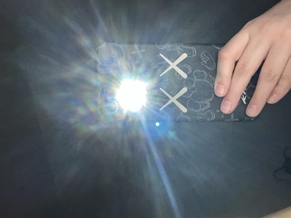 The image displays a close-up, low-light scene featuring a smartphone held by a human left hand. The smartphone, positioned vertically, has a dark-colored case adorned with white KAWS-style "XX" eyes and scattered outline graphics of KAWS Companion elements, including hands and abstract shapes. The phone's rear camera flash is actively emitting a bright white light, generating a prominent starburst effect and lens flare that dominates the upper center of the frame. A distinct smaller blue light source is visible to the immediate left of the main flash. A pale-skinned human left hand grips the lower right side of the phone, with the tips of the fingers and natural, unpolished nails visible. The background consists of an undifferentiated dark, flat surface, such as a table or desk. No identifiable landmarks or distinguishing features are present within the image to determine a specific location within the city of Izhevsk, Russia.