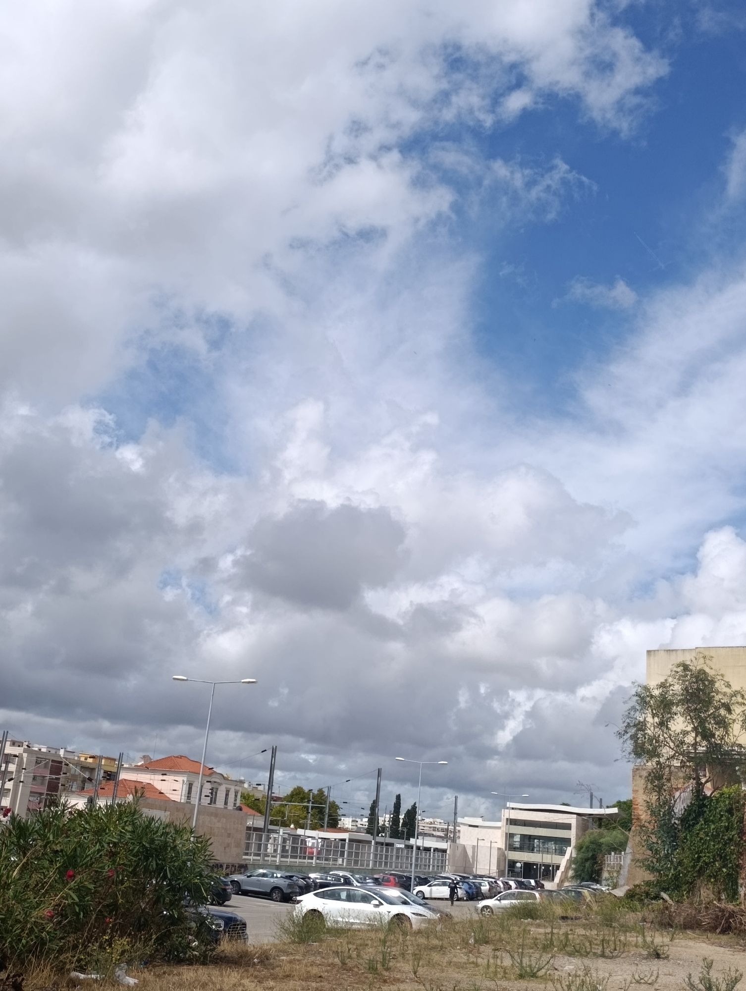 This is a photograph taken from a street-level perspective in Setúbal, Portugal, showing a parking lot filled with cars and a cloudy sky. In the foreground, dry grass and bushes are visible. A street light with two lamps stands tall, casting light. The middle ground of the image features a parking lot with various vehicles, including a white Tesla and several dark-colored cars. Behind the parking lot, buildings of different architectural styles are present. One building has a red-tiled roof and beige walls, while another appears to be a modern structure with glass elements. The sky is a mix of dark gray clouds and patches of bright blue sky, suggesting dynamic weather conditions. There are no people visibly interacting in the scene. The scene is characterized by urban infrastructure, including the street lights and buildings, combined with natural elements like the sky and vegetation.