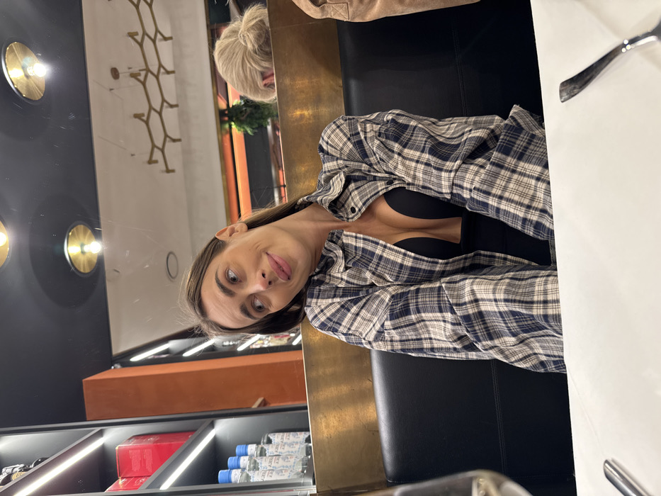 The image depicts an indoor scene within a restaurant or bar located in Verona, Italy. A woman with long brown hair, wearing an open blue, white, and beige plaid shirt over a deep V-neck black top, is seated at a table. Her head is tilted to her right, and her gaze is directed towards her left, off-camera, with a subtle smile on her lips. A white tabletop is visible in the foreground, featuring a partially obscured wine glass containing a clear liquid and a silver fork. She is seated on a black upholstered booth. In the background to her left, a dark shelving unit illuminated by LED strips displays several clear bottles with white labels and red boxes. To her right, a golden partition separates seating areas, and behind it, the upper portion of a person with blonde hair styled in a bun is visible. The ceiling above features multiple circular light fixtures with exposed bulbs and a larger, hexagonal-patterned chandelier, all contributing to the artificial, warm lighting of the establishment.