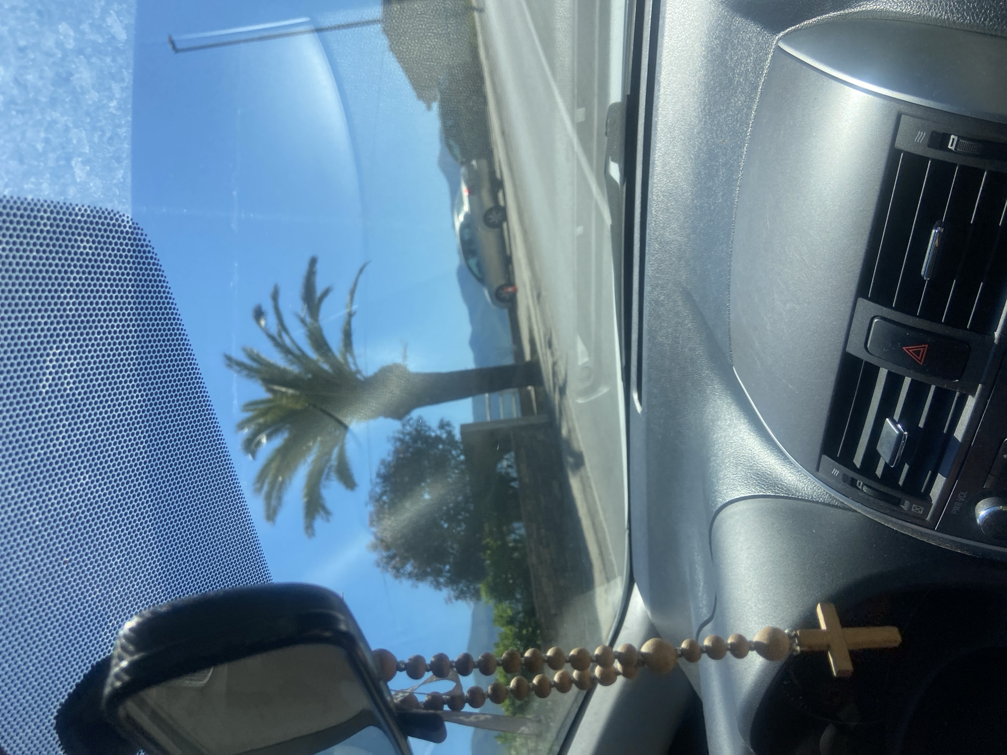 The image, taken from inside a car, shows a view of Budva, Montenegro. The car's dashboard and a wooden rosary hanging from the rearview mirror are in the foreground. Through the windshield, a palm tree is visible on the roadside, along with other greenery and a glimpse of the mountains in the background. A road runs parallel to the vegetation, with a car parked or driving on it. The sky is clear and blue.