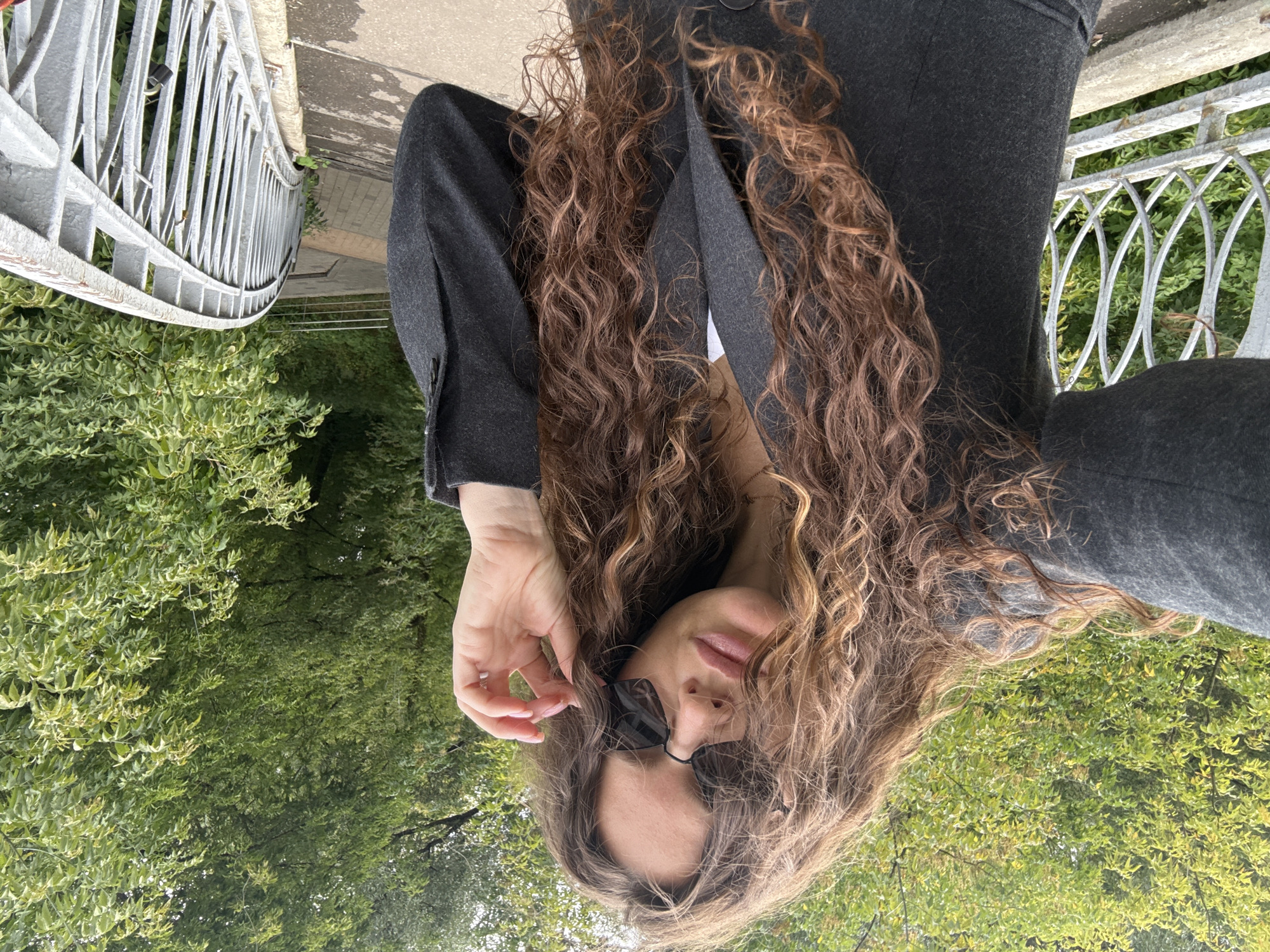 In Moscow, Russia, a young woman with long, curly brown hair and sunglasses is standing on a bridge. She is wearing a dark grey blazer and appears to be taking a selfie. Her hand is raised to adjust her sunglasses. The bridge has a white metal railing and is surrounded by lush green trees. The ground is paved with concrete and brick. A small lock is visible on the bridge railing in the background.