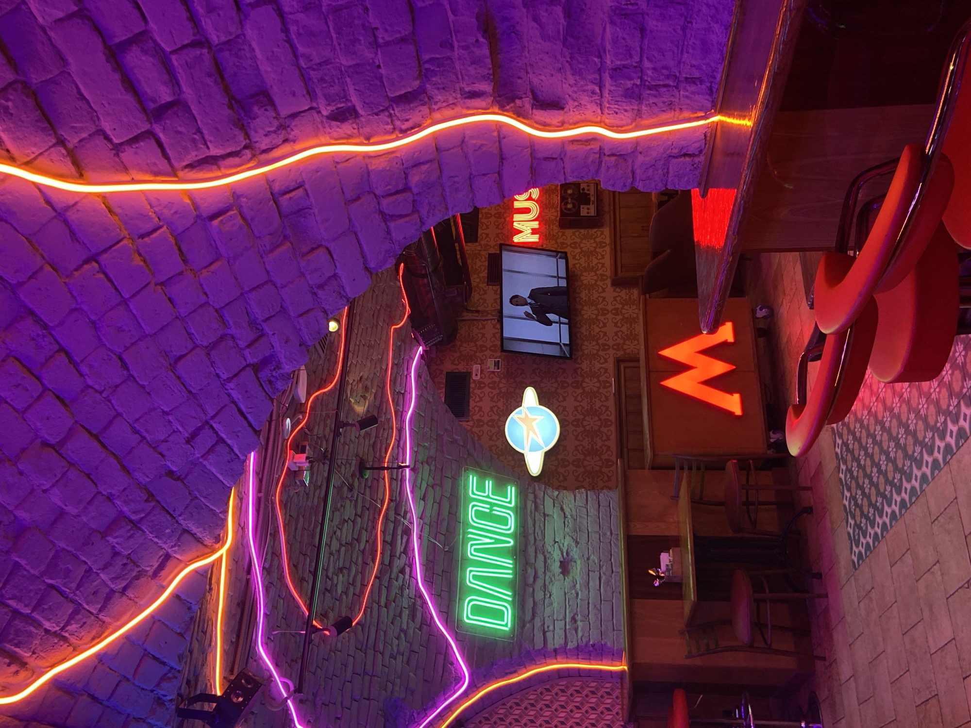 This image captures the vibrant, neon-lit interior of an entertainment venue, likely a bar or club, in Saratov, Russia. The scene is dominated by a vaulted ceiling constructed from purple-hued, brick-like material, adorned with flowing orange-yellow neon strips that illuminate its textured surface. Additional neon lights in purple, pink, and green trace intricate patterns along the walls and lower sections of the archway.
Several prominent neon signs are visible: a bright green sign boldly spells out "DANCE," another red sign partially shows "MUS" (likely part of "MUSIC" or similar), and a large, red capital letter, possibly an "M" or "W," stands near a seating area. A glowing blue and white star-shaped emblem is also affixed to a wall.
The walls feature a mix of patterned wallpaper and textured finishes, contributing to the eclectic decor. A flat-screen television is mounted on a wall, displaying a person in a suit, possibly giving a presentation or performing. Below, red-cushioned chairs or bar stools are arranged around wooden tables, suggesting a lounge or dining area. The floor is tiled with a combination of plain and patterned sections.
The overall atmosphere is lively and modern, created by the interplay of diverse lighting and textures. The absence of natural light, combined with the artificial glow, indicates it is either nighttime or an environment designed to be dark, enhancing the effect of the neon lights. No people are visible, but the setup clearly indicates a space designed for socializing, entertainment, and possibly dancing.