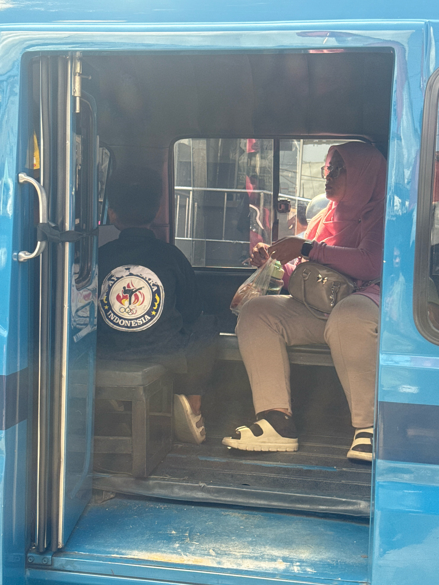 A rear view of a person wearing a black jacket with a circular emblem is visible in the back left of a blue tuk-tuk. The emblem features Olympic rings, a silhouette of a person in a martial arts pose, and the word "INDONESIA." In the front right of the tuk-tuk, a woman wearing a pink hijab and glasses is seated, holding a clear plastic bag containing a green bottle. She is wearing light-colored pants and white and black sandals. The interior of the tuk-tuk appears worn. The tuk-tuk is situated outdoors, likely in a public area, with reflections of buildings visible in the windows. The overall setting suggests a common mode of transportation in Sukaraja, Indonesia.