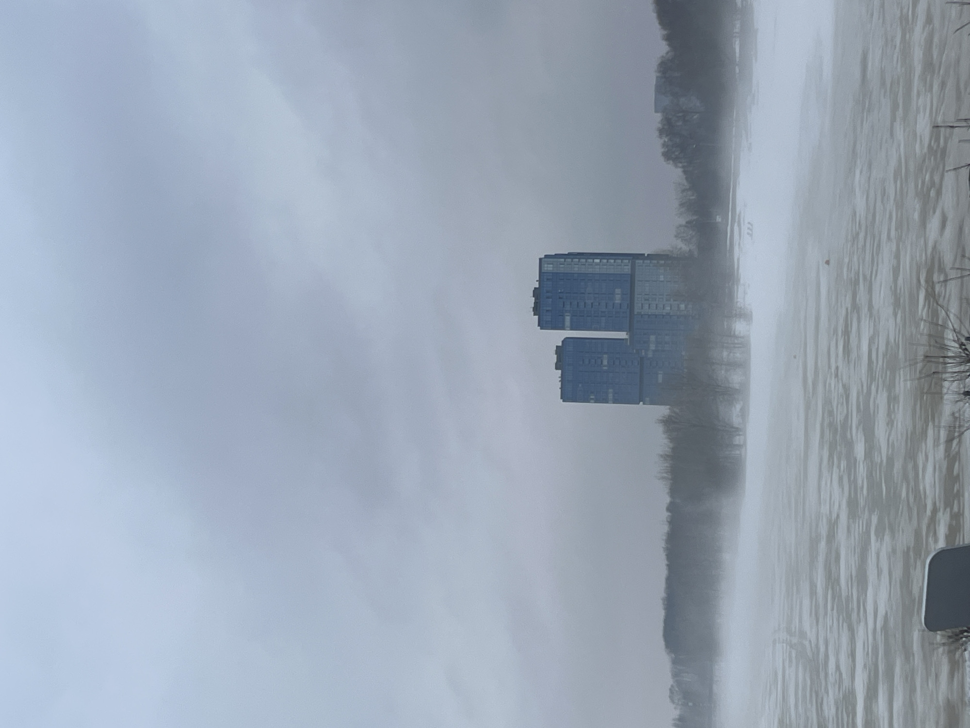 The image shows a cityscape in Kazan, Russia, on a gloomy day with a river in the foreground and a building complex in the middle ground. The building complex consists of two rectangular blue structures. A line of trees sits between the building complex and the river. The scene conveys a sense of dreariness.