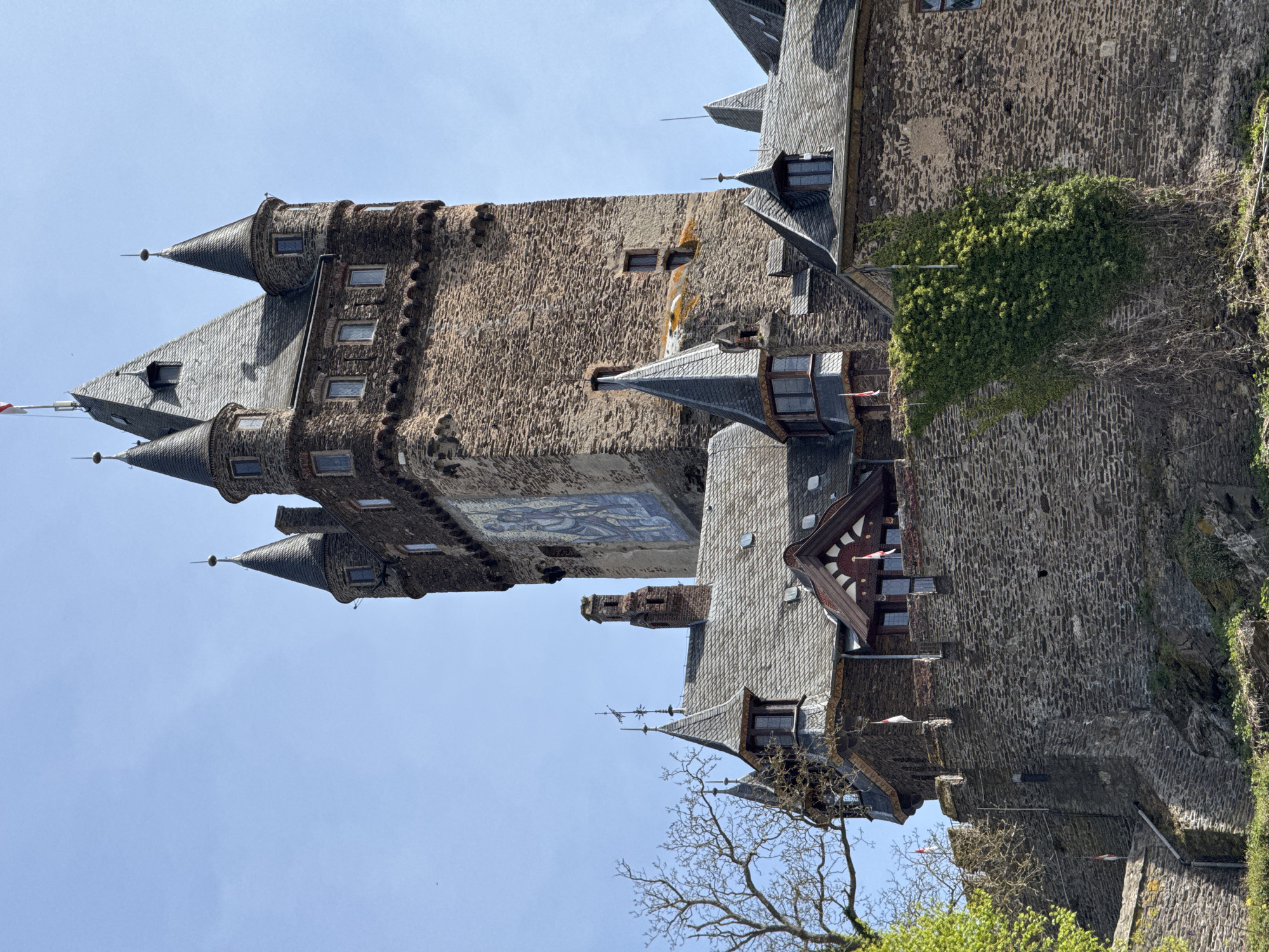 The image shows a tall, stone castle in Cochem, Germany, viewed from a low angle. The castle features multiple towers with conical roofs, small windows, and detailed masonry. One tower displays a large, colorful mural. Sections of the castle are covered with greenery, including ivy and tree branches. The sky is clear and blue.