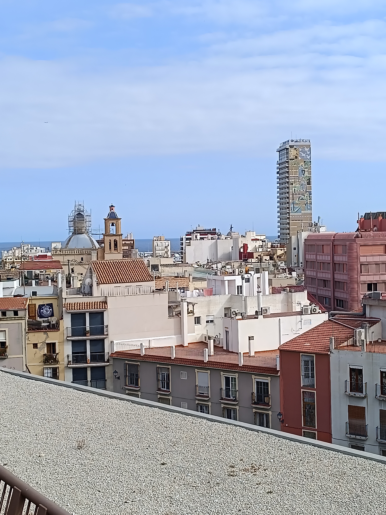 An eye-level, medium shot captures a city skyline under a bright, partially cloudy sky. In the foreground, a gravel-covered rooftop slopes down, partially obscuring a view of buildings. Beyond this, a dense collection of buildings, primarily residential, fills the frame. These structures vary in height and architectural style, with many featuring tiled roofs and balconies. A prominent feature is a tall, slender skyscraper adorned with a colorful mural, which stands out against the surrounding urban landscape. To the left of the skyscraper, a church with a dome and bell tower is visible, with scaffolding suggesting ongoing construction or renovation work. In the distance, the clear blue ocean stretches towards the horizon, indicating proximity to the coast. The scene is devoid of people, emphasizing the urban environment itself.