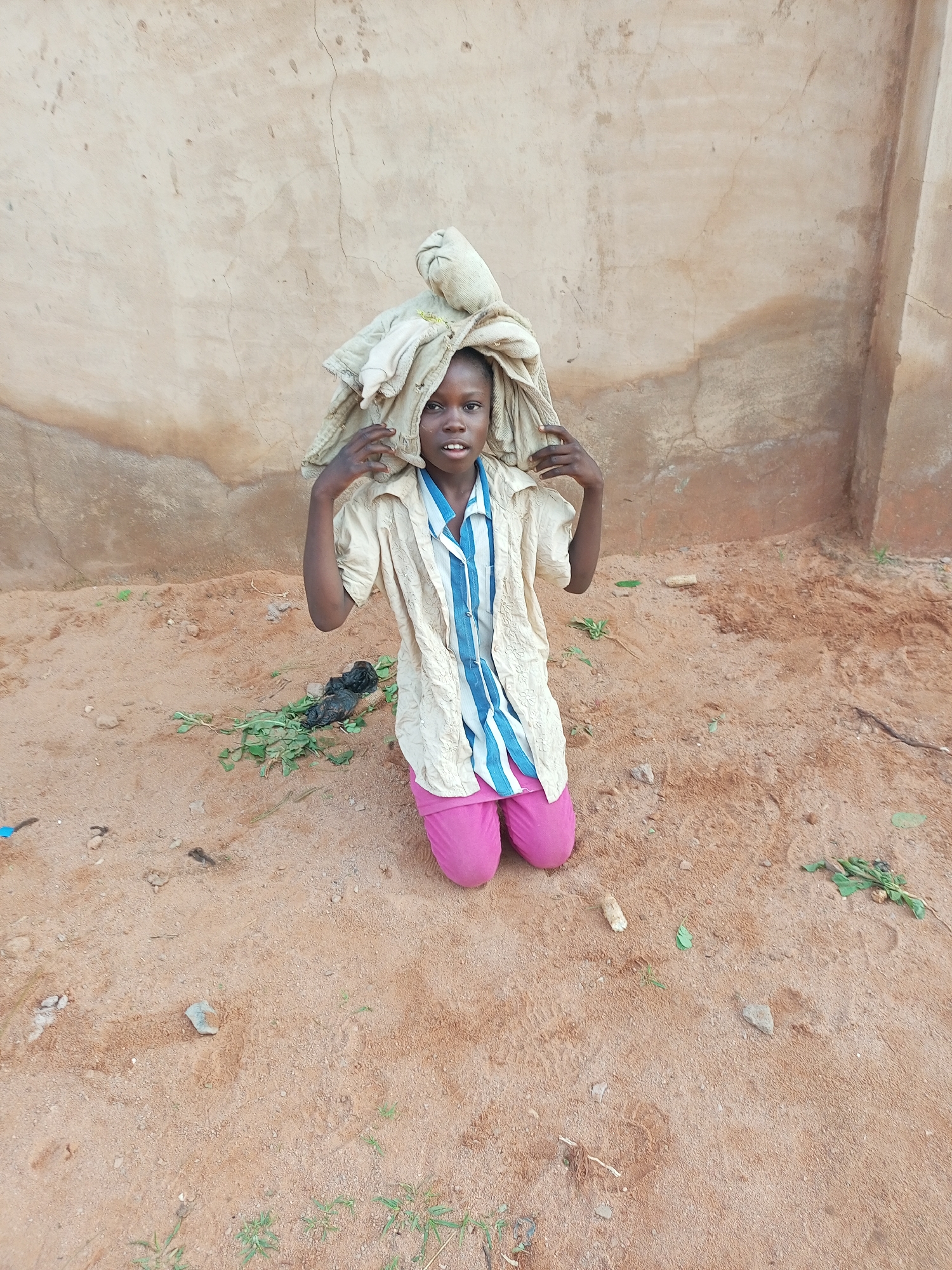 A young girl, likely of Nigerian descent and around ten years old, is kneeling on sandy ground in front of a textured, light brown wall. The wall appears weathered, with visible cracks and patches of discoloration. The girl is wearing a light-colored, short-sleeved button-up shirt with thin blue stripes and bright pink trousers. On her head, she balances a large bundle of light-colored fabric, possibly laundry. Her hands are placed on either side of the bundle, adjusting its position. The ground is a mix of sand and small stones, with sparse green vegetation scattered around. A black plastic bag and some leaves are also visible on the ground near the girl. The lighting suggests it is daytime, with soft shadows indicating an overcast sky or indirect sunlight. The overall impression is one of a child performing a chore, possibly carrying goods, in a dry, rural or semi-urban environment within Tempure, Nigeria.