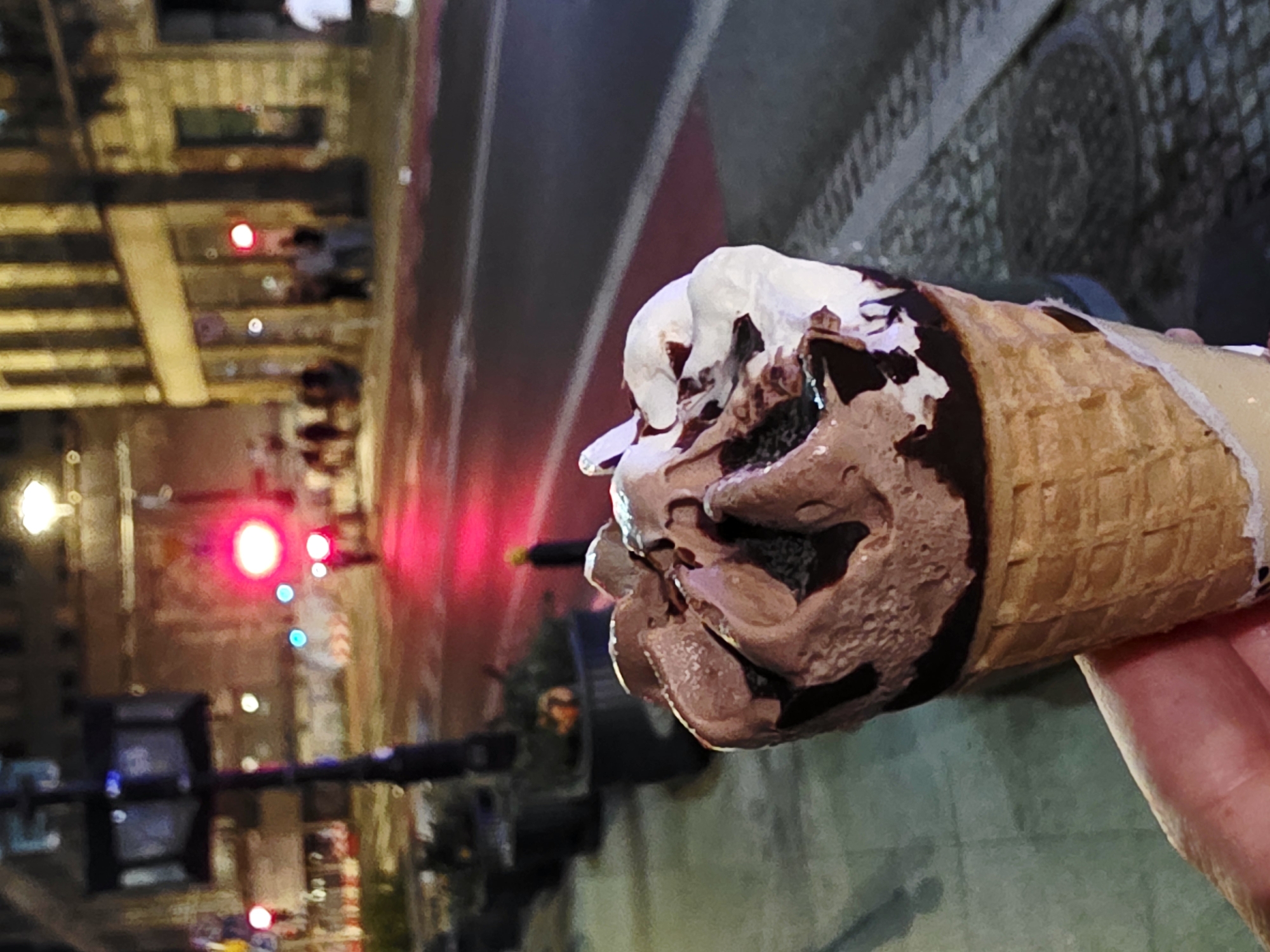 A close-up, shallow depth-of-field shot shows a chocolate and vanilla ice cream cone held in a person's left hand in the foreground. The cone is a waffle cone and the ice cream has drizzles of chocolate sauce. In the background, a street scene is visible at night. Red and white lights from traffic signals and streetlights illuminate the street, which appears wet. Buildings line the street on the right side. A few blurred figures of people are visible on the sidewalk. The overall impression is of an urban setting at night, possibly in Warsaw, Poland, given the architecture and street lighting.