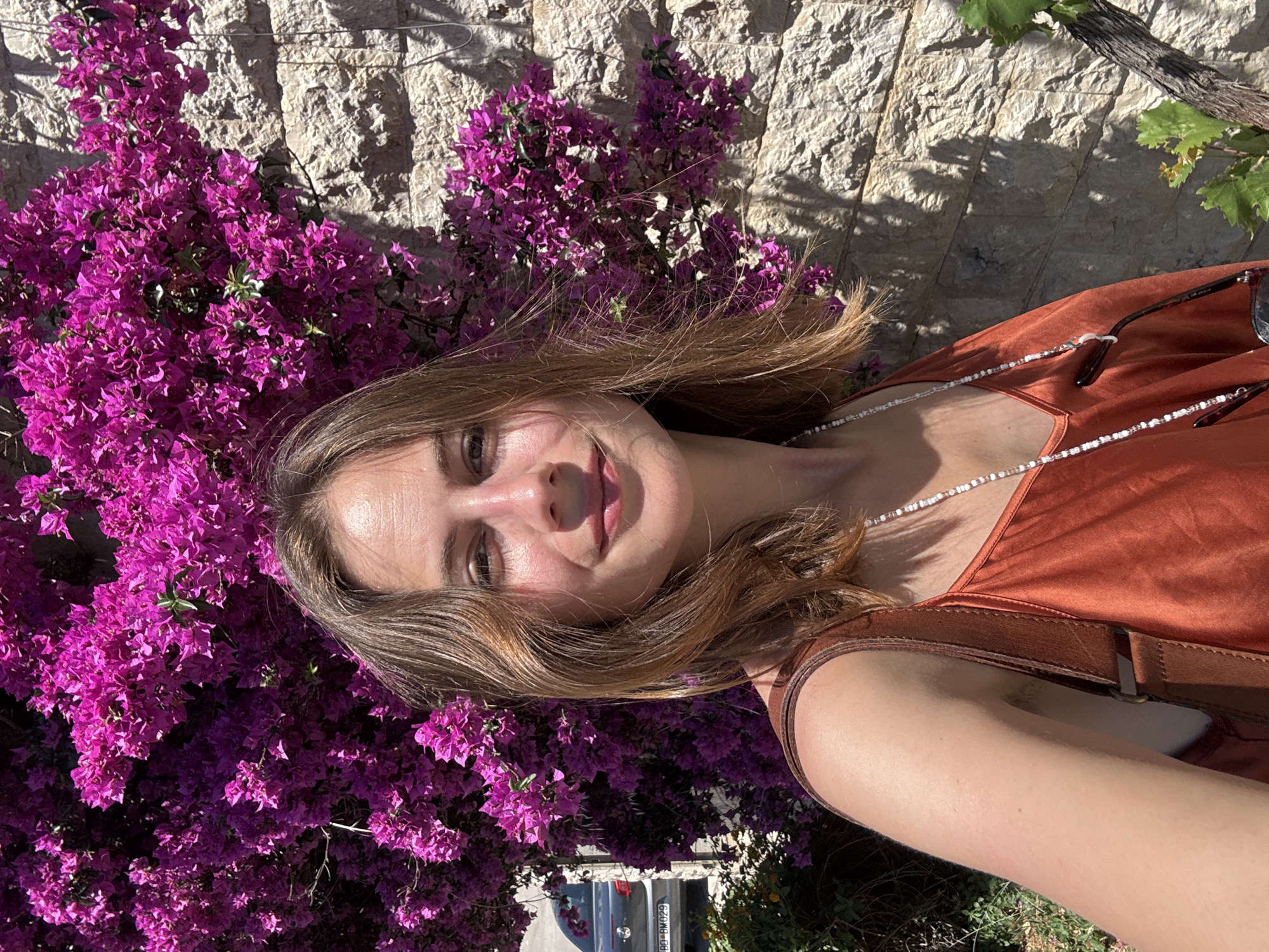 A young woman stands in front of a wall in Boreti, Montenegro, with a large bush of vibrant pink bougainvillea flowers to her left. She is smiling and looking directly at the camera, holding her phone to take a selfie. She is wearing a rust-colored, sleeveless dress with a thin strap over her shoulder and a beaded necklace. Sunglasses are attached to the necklace and rest against her chest. Behind her to the left, a dark-colored car is visible, partially obscured by the bougainvillea. The wall behind her is made of rough, light-colored stones, and sunlight casts shadows from the bougainvillea and a nearby vine onto the wall.