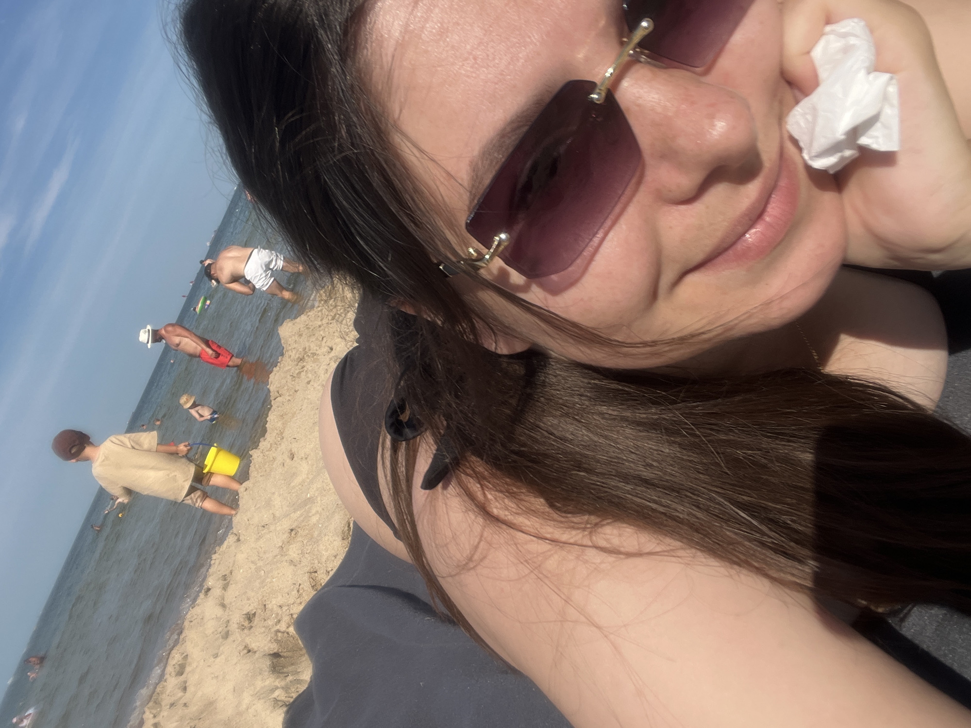 The image depicts a sunny day at a beach, likely in the area of Gdańsk, Poland. In the foreground, a person is lying down, with their face, shoulder, and arm visible. They are wearing sunglasses and have dark hair. A crumpled white tissue is held in their hand. In the background, other beachgoers are visible, including children playing in the shallow water, one with a yellow bucket. Adults are also seen wading and standing in the sea, with a few more in the distance. The environment is characterized by sand, water, and a clear blue sky with some clouds.