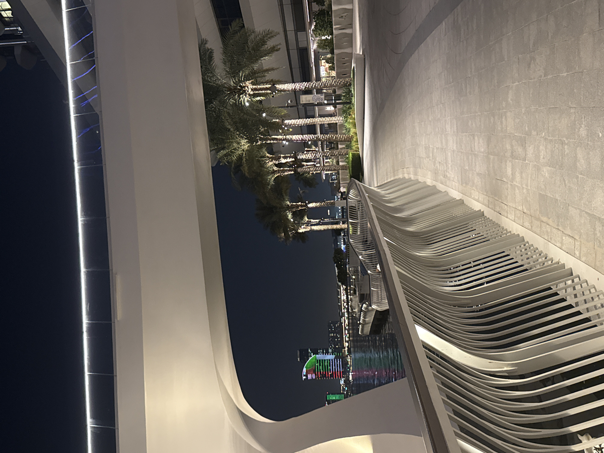 The image, taken at night, shows an architectural structure with palm trees and a skyline featuring a lit-up building in the city of Dubai, United Arab Emirates.
The foreground is dominated by a curved white structure, possibly part of a modern building or an artistic installation. This structure frames the view beyond. Adjacent to the structure, there are several rows of curving white metal slats.
Through the opening created by the structure, a row of palm trees extends into the distance, partially obscuring the skyline. The trees are lit, casting a soft glow on their fronds.
The skyline is punctuated by illuminated buildings. The focal point is a distinct structure adorned with vertical stripes of red, white, and green lights, evocative of a national flag. Further back, other buildings contribute to the skyline, adding to the urban ambiance. The reflection of the lights shimmers on what appears to be water.
The scene captures a blend of modern architecture, natural elements, and urban lighting, reflecting Dubai's distinctive cityscape.