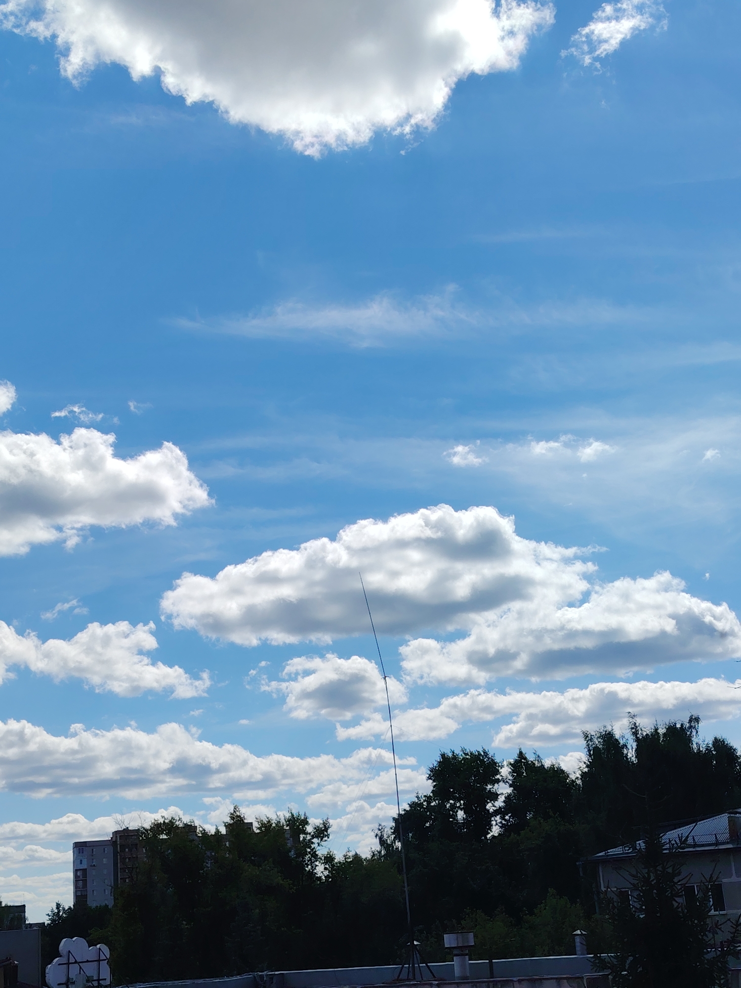 This is an outdoor, eye-level, vertical photograph taken from what appears to be a rooftop in the city of Konakovo, Russia. The sky dominates the upper portion of the image, filled with a vibrant blue hue and a scattering of white, puffy cumulus clouds. The clouds vary in size and density, with some appearing bright and sunlit, while others are shaded.
The lower part of the image shows a cityscape with a dense line of dark green trees forming a backdrop. Behind the trees, a multi-story residential building with a light-colored facade is visible. To the right, another building with a sloped, gray roof and some visible windows is present. In the foreground, a concrete structure with some white objects is partially visible.
A tall, thin, dark antenna or pole extends vertically from the bottom left of the frame into the sky, disappearing among the clouds. The overall impression is a sunny day with a partly cloudy sky over a residential area. There are no people visible in the image.