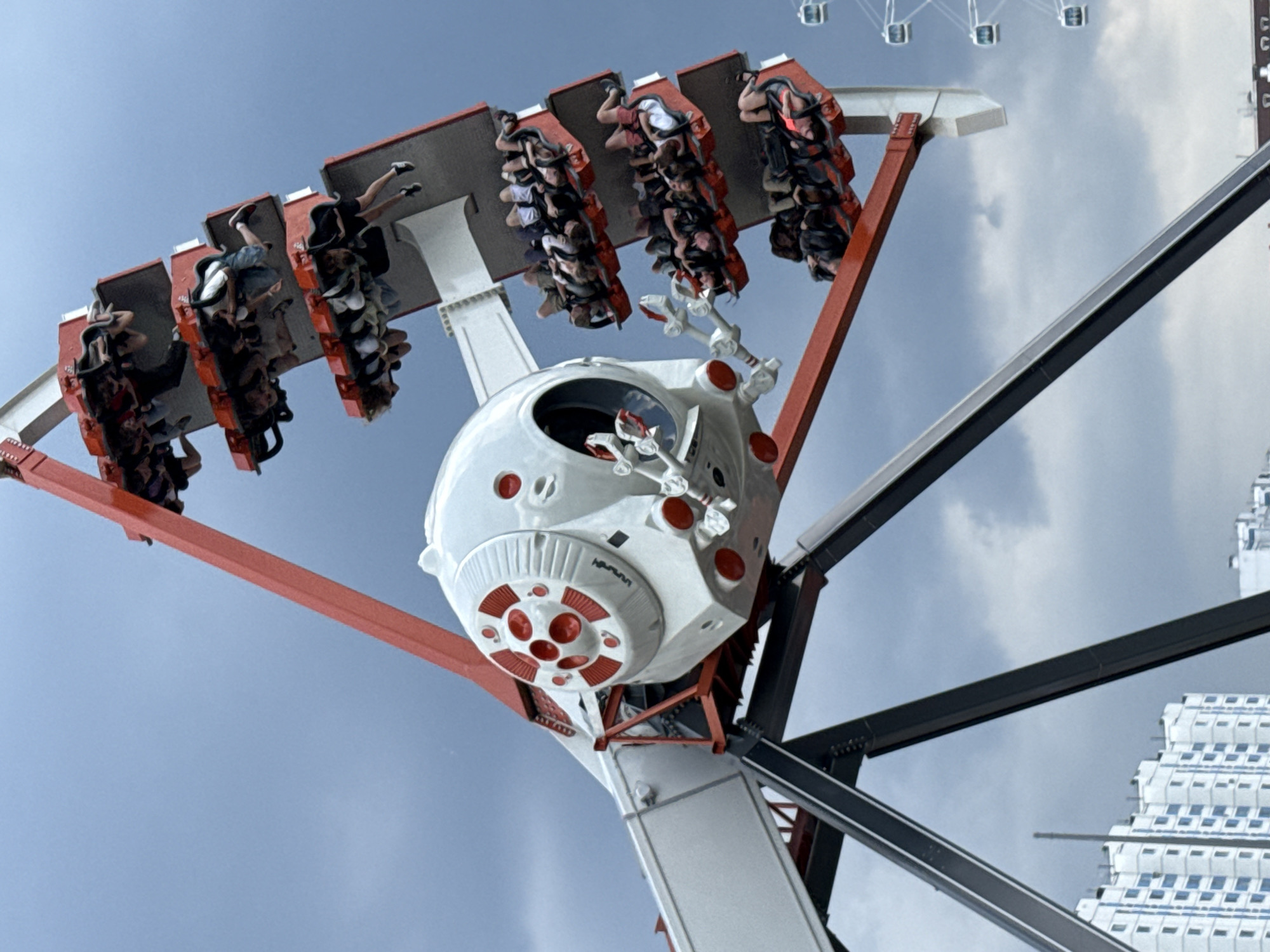 In Khimki, Russia, an amusement park attraction, likely a pendulum ride, is in motion against a cloudy sky. The ride features a central white and red pod-like structure with several arms extending outwards, each holding a row of passenger cars. At least four rows of cars are visible, each filled with people who are hanging upside down or at steep angles due to the ride's movement. The structure is supported by large, dark metal beams. In the background, a Ferris wheel with multiple gondolas and a tall residential building with blue-trimmed windows can be seen, indicating an urban setting. The overall scene captures a moment of thrill and excitement at an amusement park.