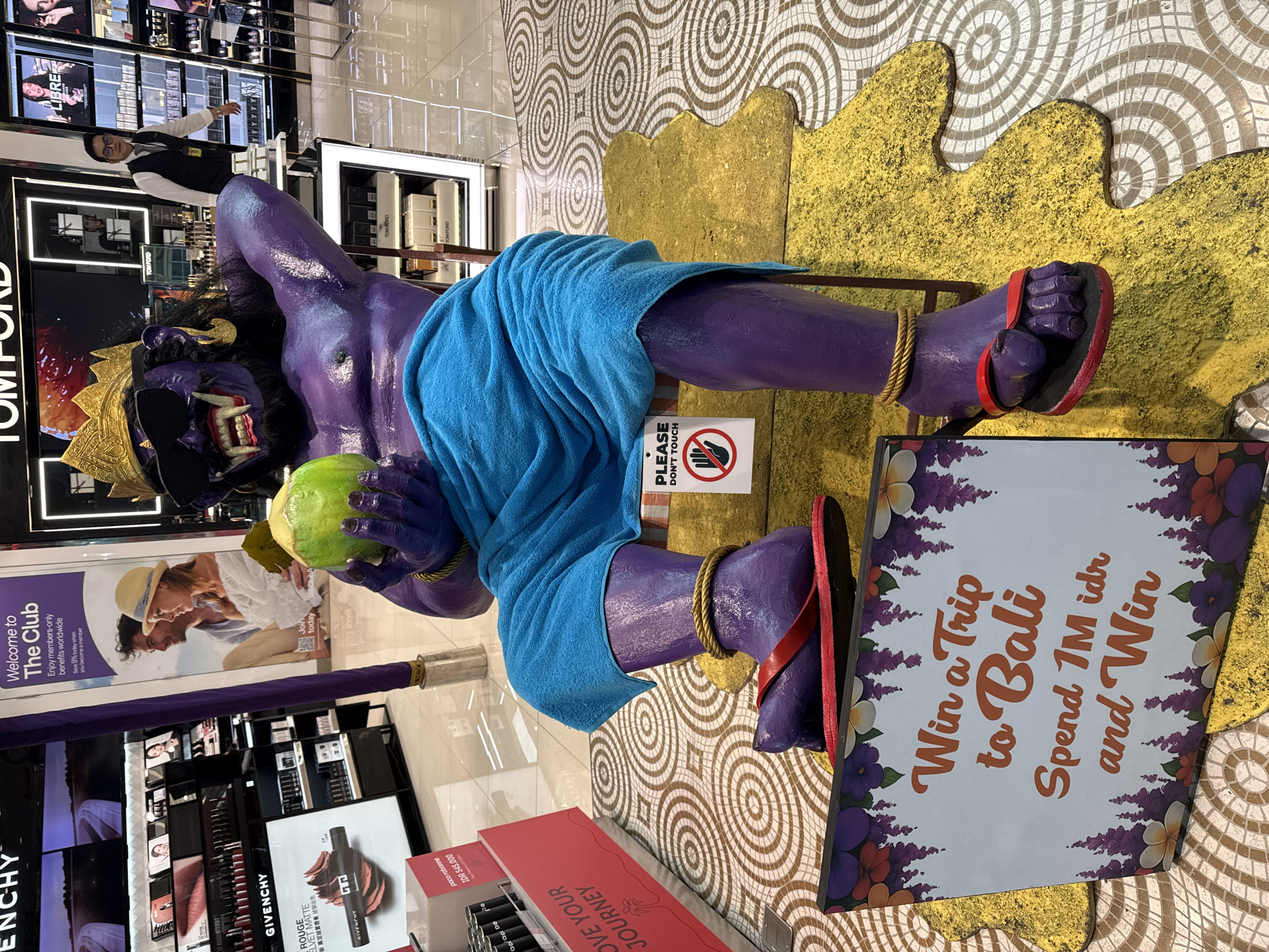 A purple, human-like statue wearing a gold crown, sunglasses, and a blue cloth is seated on a chair in what appears to be a high-end duty-free shop. The statue is holding a green coconut in its left hand and has a golden rope tied around its ankles and wrists. A sign in front of the statue reads "Win a Trip to Bali, Spend 1M idr and Win". The statue is placed on a yellow, abstract-shaped mat that resembles a spilled liquid. The background shows displays of makeup and perfume products from brands like Givenchy and Tom Ford, indicating a retail environment. A store employee in a uniform is visible in the background. The floor is tiled with a circular pattern. The location is identified as Kuta, Indonesia, a popular tourist destination known for its beaches and shopping.
