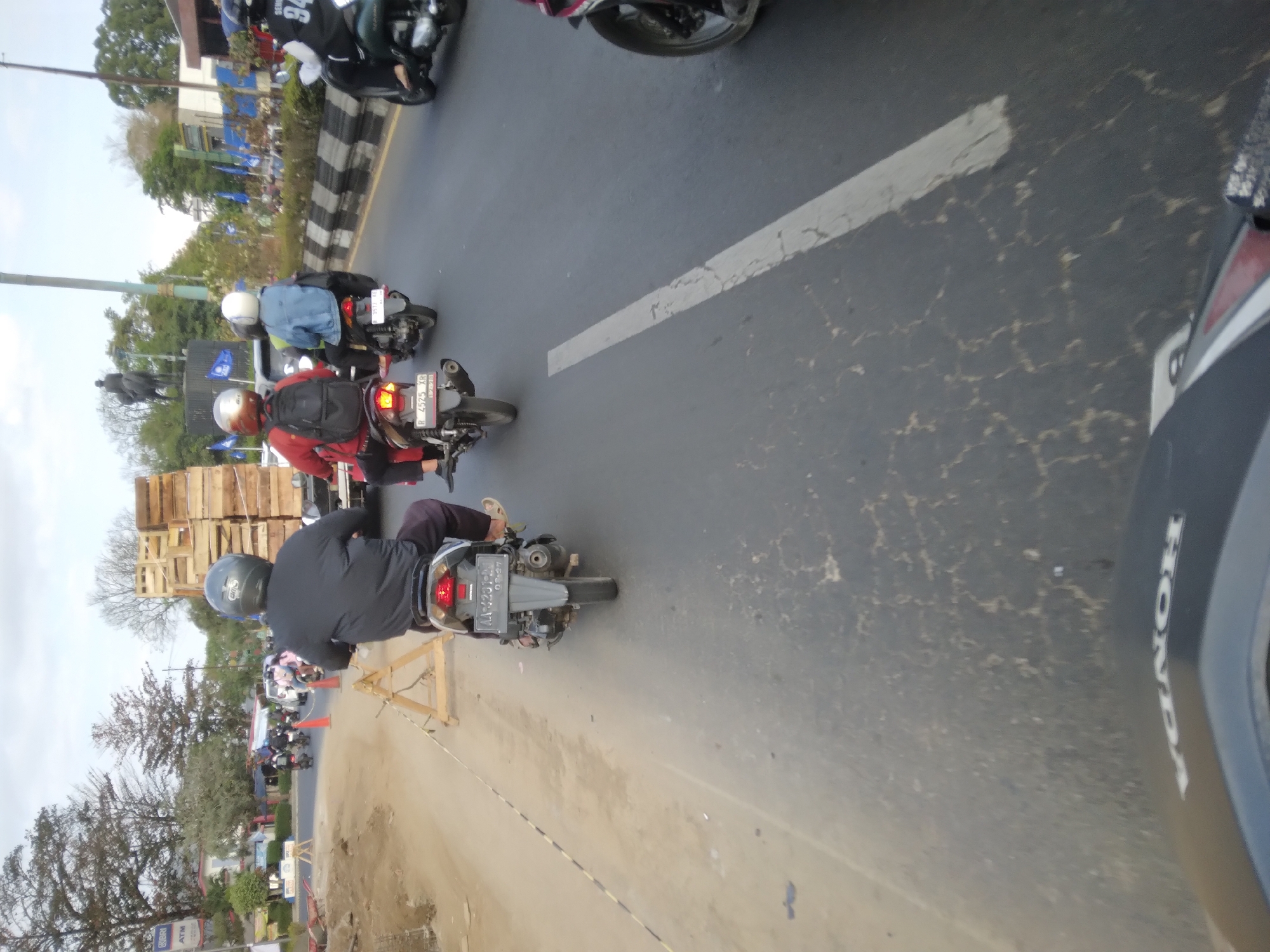 In Karangnanas, Indonesia, a first-person perspective shot captures a street scene with multiple motorcycles navigating a road. The immediate foreground shows a portion of a black motorcycle's front, with the "HONDA" logo visible. Ahead, a rider on a motorcycle wearing a black jacket and grey helmet is in focus. Behind them, a second motorcycle carries a rider in a red jacket and black helmet, with a backpack. Further down the road, a third motorcycle is ridden by a person in a blue jacket and white helmet. The road itself is paved and appears somewhat worn, with a white line marking the lane. To the left, a construction area is visible with dirt and yellow caution tape. To the right, a roadside is lined with a black and white striped barrier, trees, and buildings. Several blue flags with white emblems are planted along the roadside. In the background, a statue of a person on horseback stands prominently. The sky is overcast.