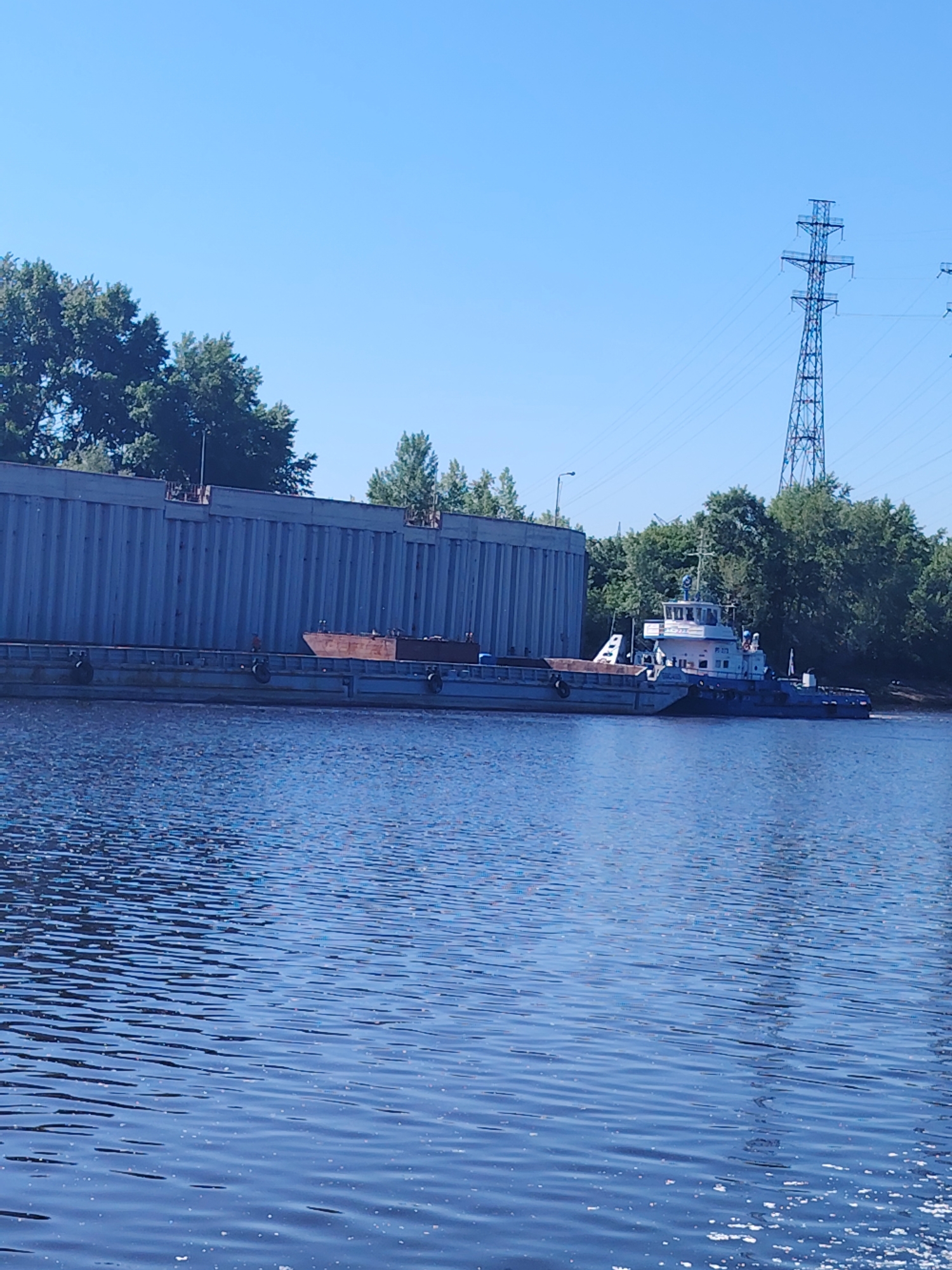 A wide, clear blue sky is visible above a calm body of water, likely a river or canal, in Chaikovsky, Russia. On the water, a long, gray barge is moored alongside a large, grey, ribbed structure, possibly a seawall or a building under construction. The barge appears to be carrying some reddish-brown cargo, possibly metal or construction materials. A blue and white tugboat is attached to the rear of the barge. In the background, a line of green trees forms a natural border, with a tall, metallic power pylon visible on the right side of the image. The water surface shows gentle ripples and some scattered white foam or debris.