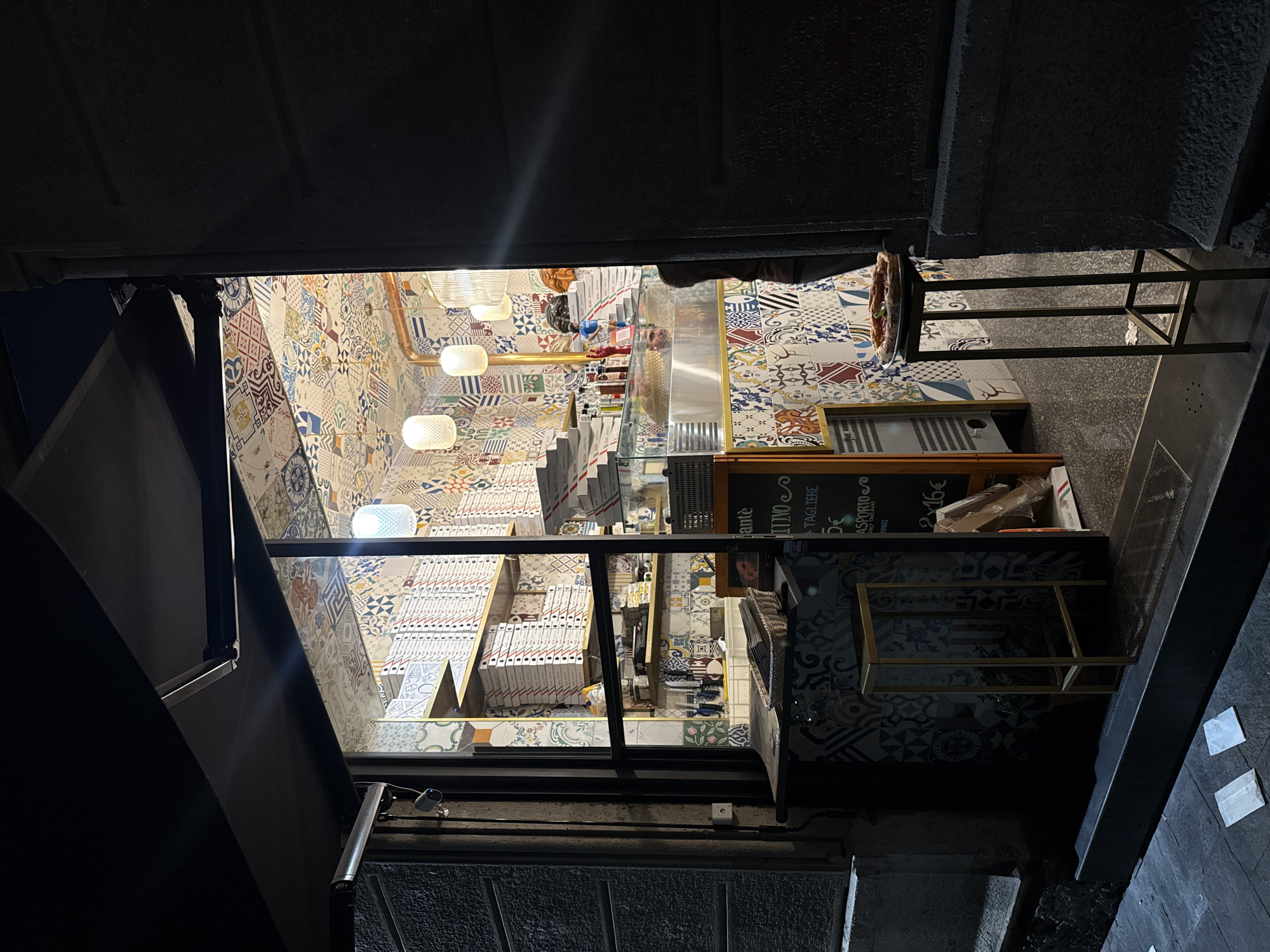 The image depicts the interior of a pizza restaurant in Milan, Italy, seen from an exterior, elevated perspective. The walls and floor are tiled with a patchwork of colorful patterns. Inside, stacks of pizza boxes are arranged on shelves. A service counter and preparation area are visible, illuminated by hanging lights. The view is framed by dark, architectural elements, possibly part of a bridge or building overhang, giving the impression of peering into the restaurant from a higher vantage point. The exterior is dark, suggesting it's night.