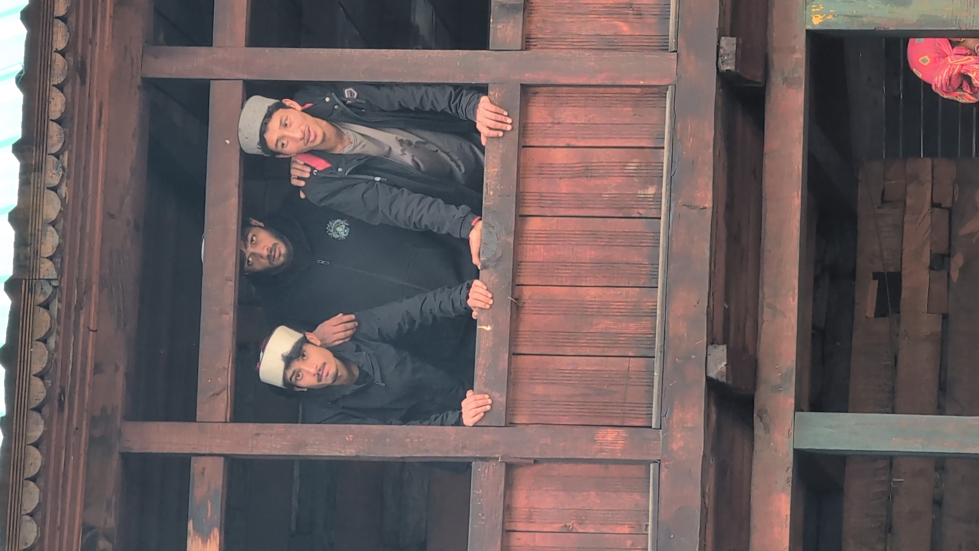 Three young men are standing on a wooden balcony in Malana, India. The man on the left wears a white and maroon cap, a black jacket, and is looking at the camera. The man in the middle wears a black hoodie and is looking slightly to the left. The man on the right wears a grey cap, a black jacket over a grey t-shirt, and is looking to his right. The balcony is made of dark wood planks and posts, and the building appears to be constructed from wood as well. In the background, another person wearing a pink headscarf is partially visible. The lighting suggests it is daytime.
