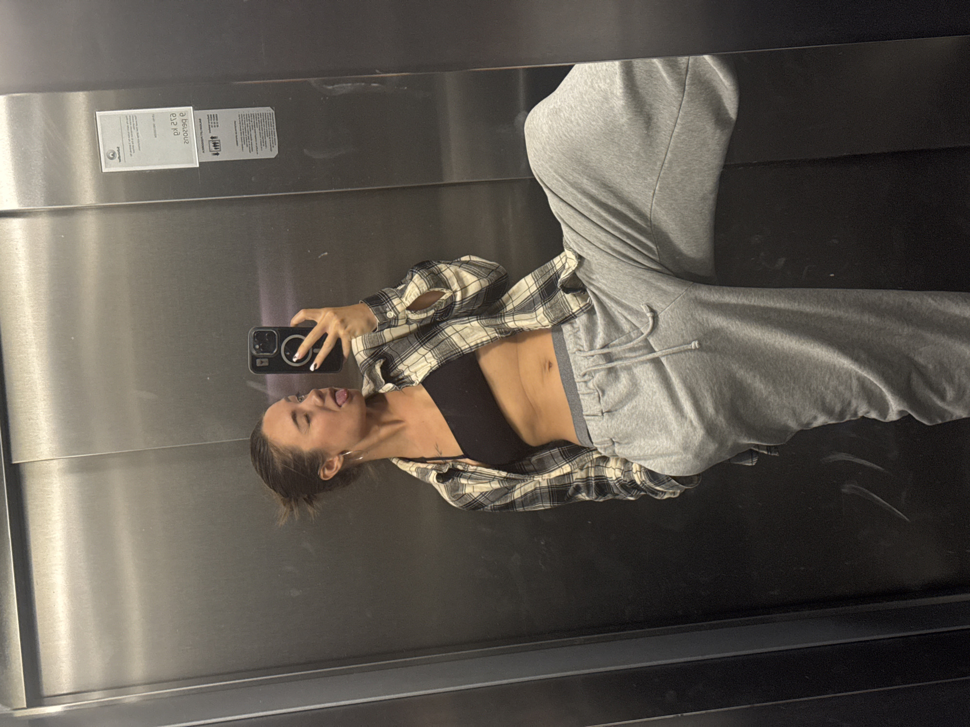 A young woman is taking a selfie in what appears to be an elevator, her reflection visible in the mirrored surface. She is wearing a black sports bra, grey sweatpants, and an open plaid flannel shirt. Her hair is pulled back in a bun.
The elevator's interior is metal, with a small information panel visible on the upper left. The location of the photo in the city of Budva, Montenegro is not discernable.