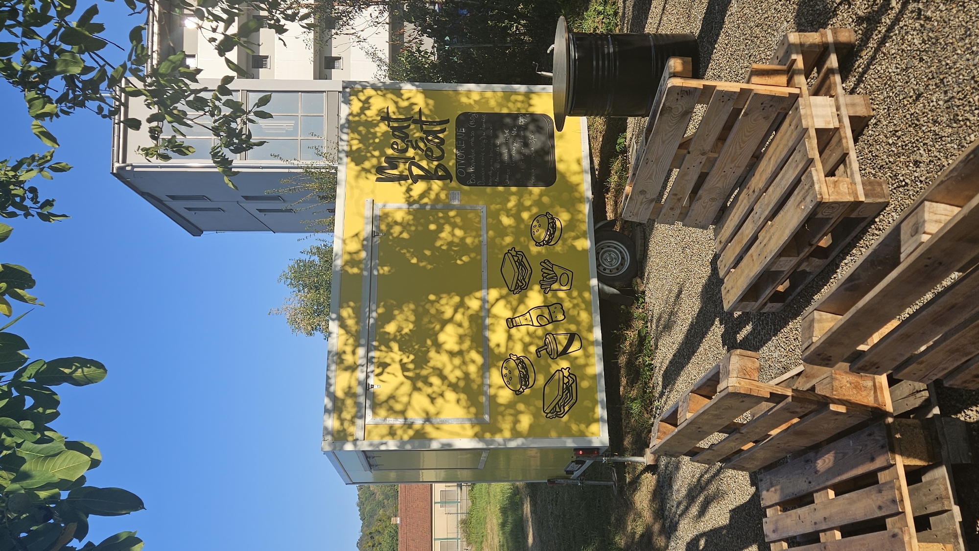 The image depicts an outdoor scene in the city of Prešov, Slovakia, featuring a vibrant yellow food truck named "Meat Beat". The food truck is adorned with cartoonish illustrations of burgers, sandwiches, a soda cup, a bottle, and french fries. A chalkboard menu is attached to the side of the truck, listing prices in Euros. In front of the food truck, several seating arrangements made from wooden pallets are visible on a gravel ground. To the right of the food truck, a black barrel-like container with a metal lid, possibly a grill or trash bin, stands. The background shows a modern apartment building, trees, and a building with a reddish roof, suggesting an urban or suburban setting. The sky is clear and blue, indicating a sunny day. There are no people visible in the image.