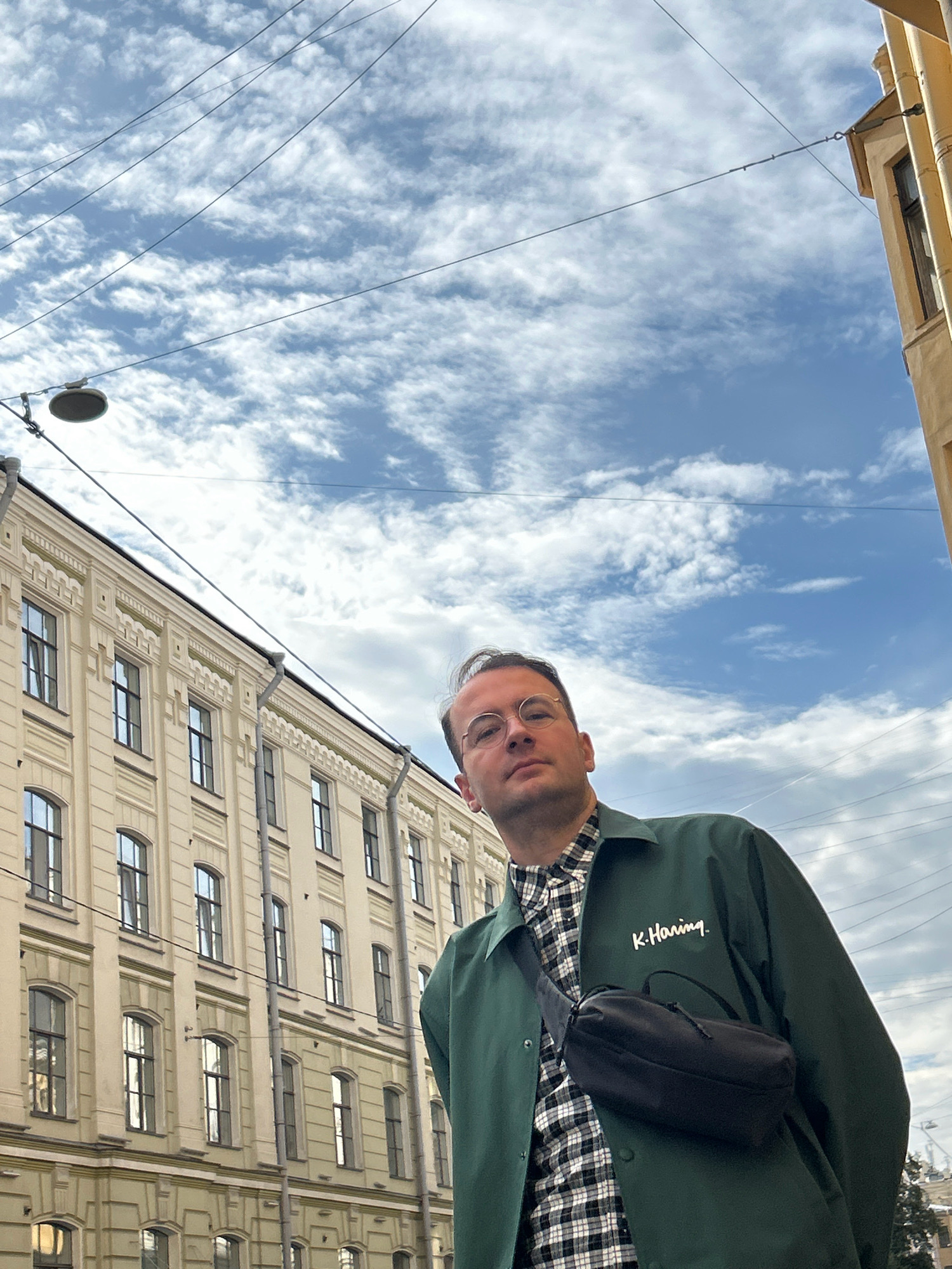 A man with short brown hair and glasses is standing outside in front of a building. He is wearing a green jacket with "K.Haring." embroidered on the left breast, over a black and white checkered shirt. A black fanny pack is slung across his chest. The building behind him is pale yellow with numerous windows and decorative molding. Overhead, a network of power lines crisscrosses a cloudy blue sky. This scene is likely in Saint Petersburg, Russia, given the architectural style of the building.