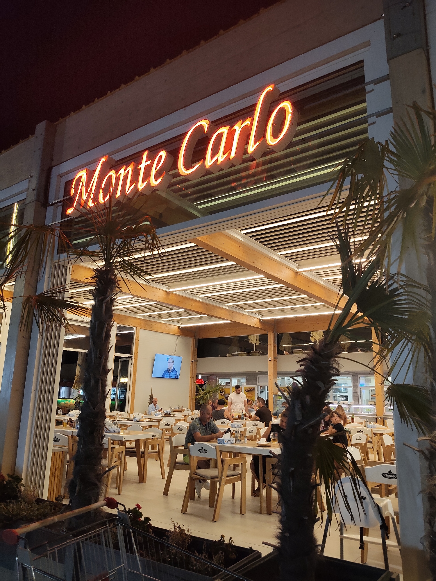 The image shows a restaurant named "Monte Carlo" with neon lights, located in Constanța, Romania. The establishment features an outdoor seating area with tables and chairs, partially covered by a wooden and slatted roof illuminated by strip lights. Several patrons are seated at the tables, engaged in conversation or dining. Palm trees are visible in the foreground and to the sides, suggesting a warm climate or a tropical theme. A television screen is mounted on a wall inside the restaurant, displaying a man's face. A shopping cart is partially visible in the lower left corner of the image. The overall atmosphere appears to be a lively evening dining experience.
