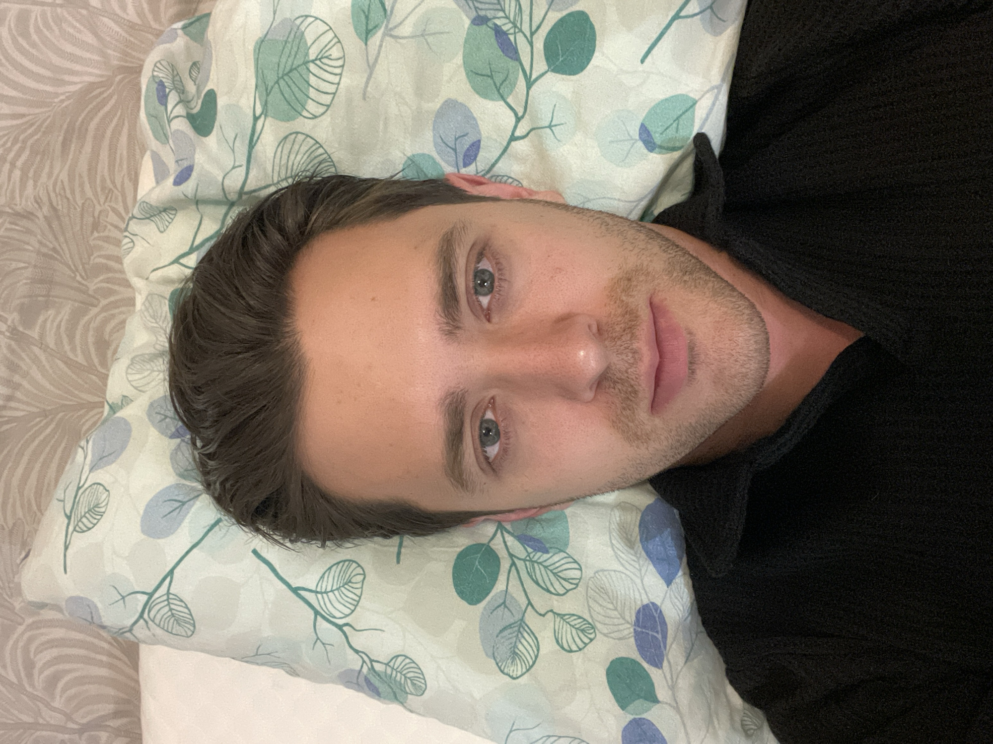A light-skinned man with dark hair lies on a pillow with a green and blue leafy pattern. He is wearing a black shirt. The pillow is on a bed with a patterned bedspread. No location is identifiable from the image.