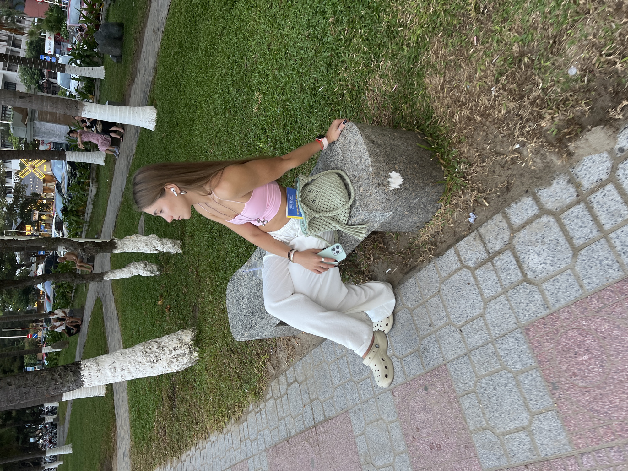 A young woman sits on a stone bench in a park in Nha Trang, Vietnam. She is facing to her left, looking away from the camera. She is wearing a pink crop top, white wide-leg pants, and beige Crocs. She has on white wireless earbuds and a silver necklace with a pendant. On her lap, she holds a light green woven bag and a smartphone. To her right, on the bench, is a blue book. The park has palm trees with white-painted bases and a paved walkway with red and gray tiles. In the background, there are cars, buildings, and other people sitting on benches. The ground is covered with grass.