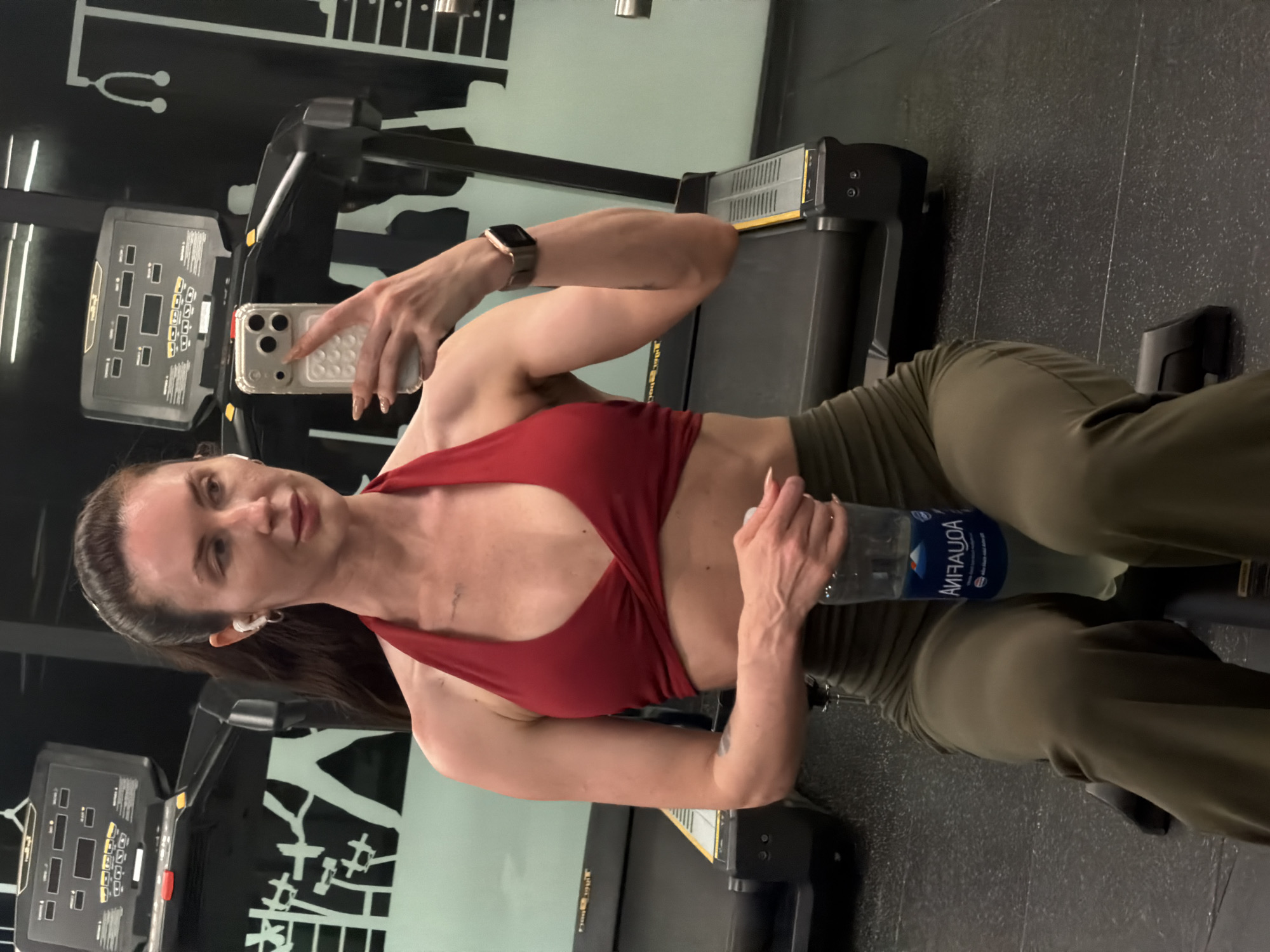 In a gymnasium in Dong Ninh Hoa Ward, Vietnam, a woman with dark hair tied back and wearing wireless earbuds, a red halter top, and olive green athletic pants sits on a bench, holding a partially filled bottle of Aquafina water. She is taking a selfie with her phone, which is held in her right hand. In the background, two treadmills are visible, with a graphic of weightlifting figures and a stethoscope on the wall behind them.