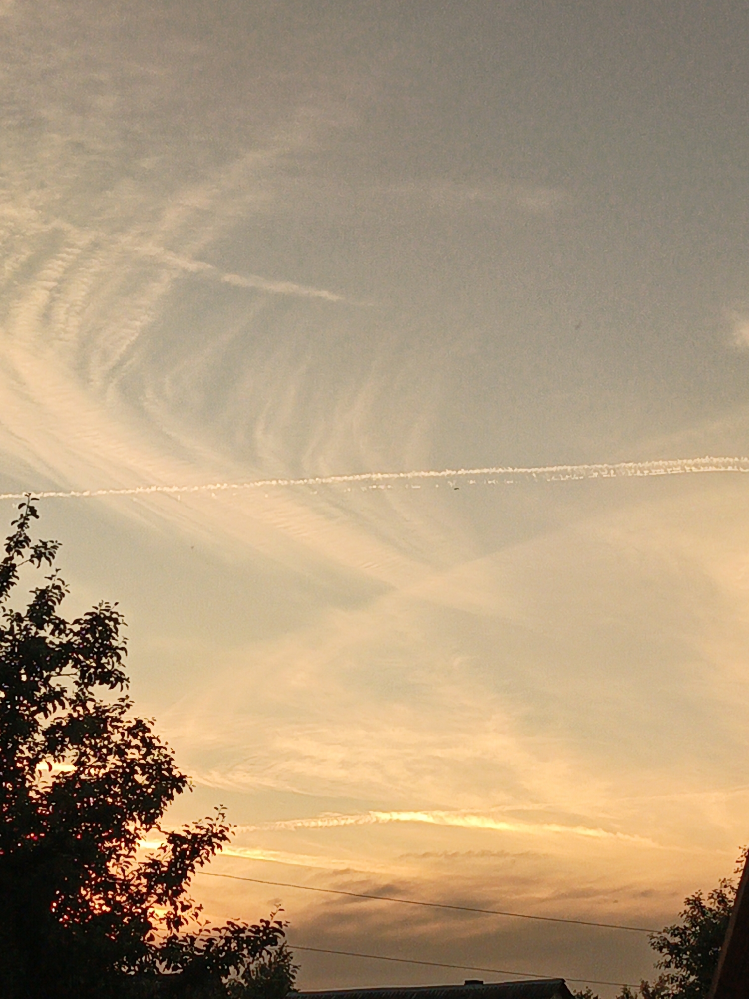 The image shows a sunset sky with clouds and contrails over a rural or suburban area. A silhouetted tree is in the foreground on the left, with the sunset light filtering through its leaves. A roofline of a building is visible at the bottom of the frame. There are power lines stretching across the scene. The sky has a gradient of colors, with lighter tones at the top transitioning to warmer oranges and yellows near the horizon.
