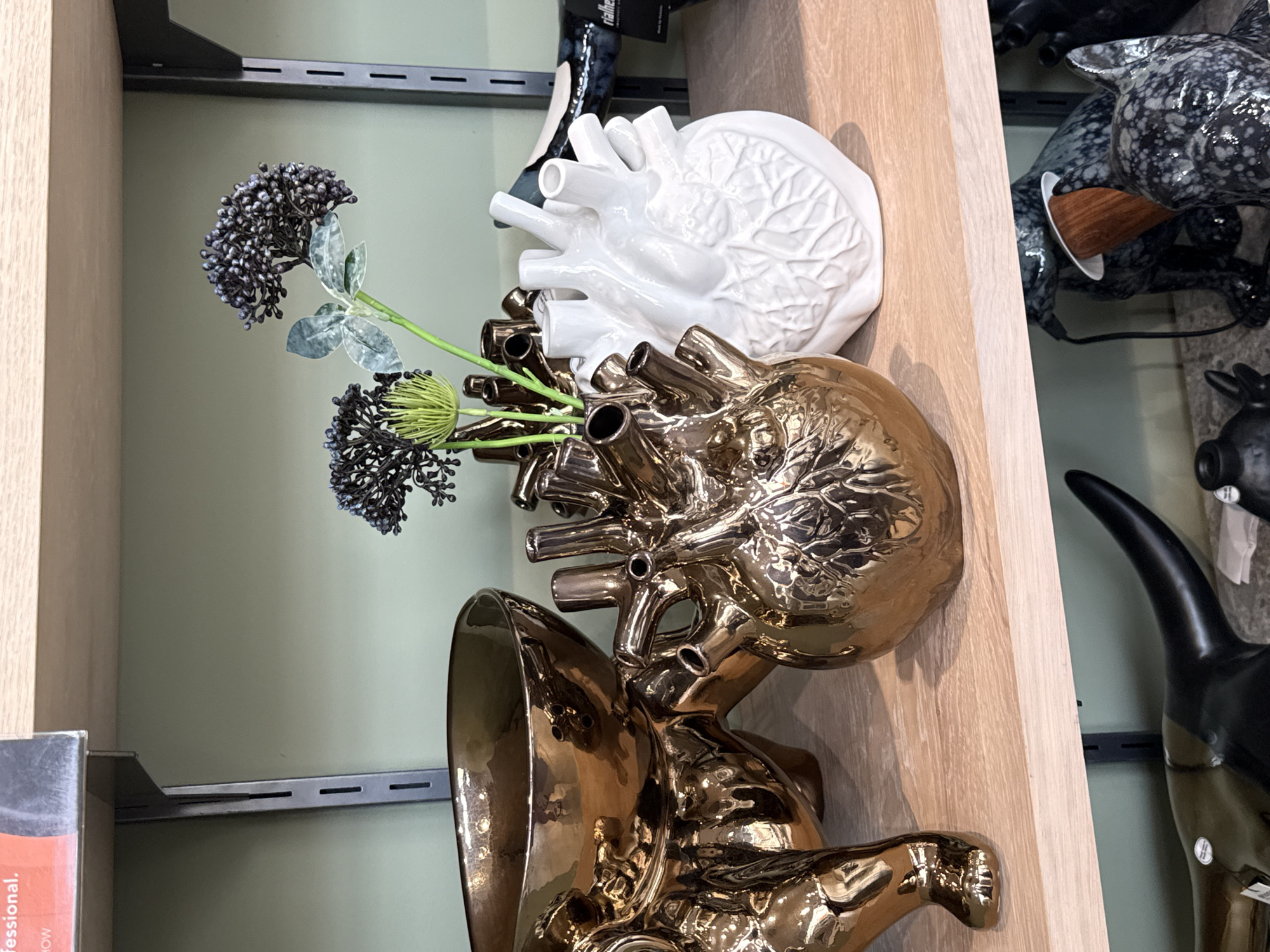 The image shows a shelf in a retail environment. The shelf, made of light-colored wood, holds a variety of decorative items. From top to bottom, there's a white ceramic heart-shaped vase, a gold-colored ceramic heart-shaped vase with dark purple flowers, and a gold-colored vase with a design that appears to be an animal skull, possibly a bull. To the right of the wooden shelf, there are additional decorative items, including a spotted sculpture and a black bull sculpture. The background features a pale green wall and shelving supports. The location details (Cape Town, South Africa) are not visually verifiable from the image content.