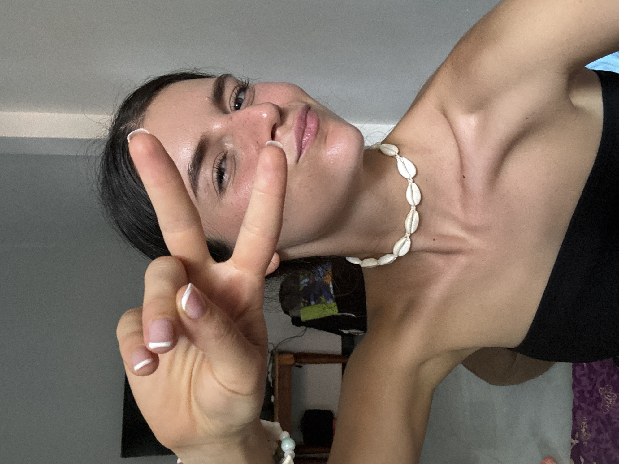 A fair-skinned woman with dark hair is the central subject of this selfie. She is positioned at an angle, tilting her head to the left and extending her right hand in front of her face, forming a peace sign. Her nails are painted with a white French tip design. She wears a necklace composed of white shells and a black sleeveless top. Her facial features include freckles, and her skin appears slightly damp.
The background reveals a corner of a room. Part of a shelf with various objects on it, including what appears to be a framed photograph, is visible. A flat-screen television is partially visible on the left side of the frame. A section of patterned fabric in shades of purple and white is visible in the lower right corner.
There is no definitive information to identify the image location as Mui Ne, Vietnam, from the visual details provided.