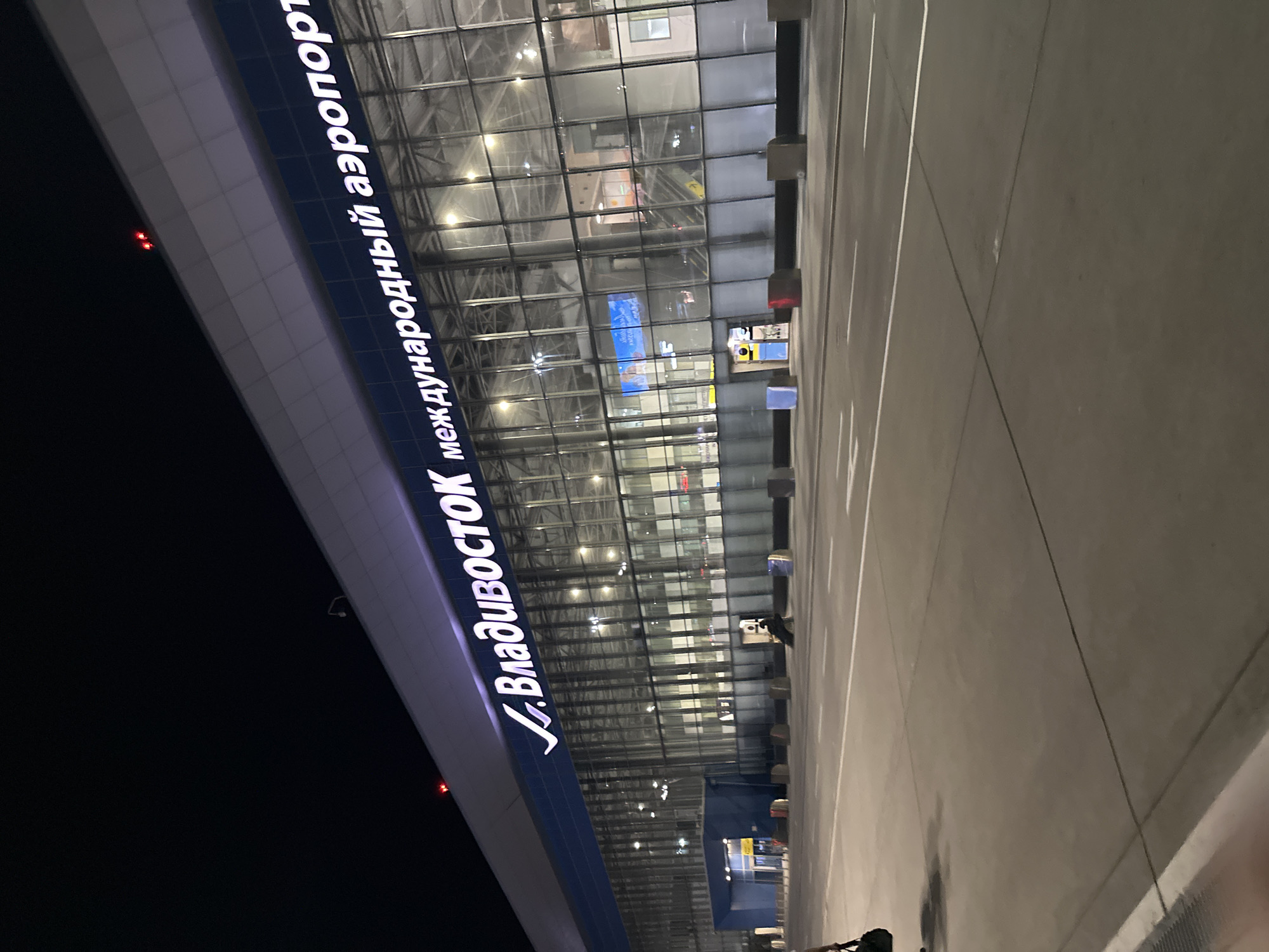 The image shows the exterior of an airport at night. The building has a modern design with a large glass facade. Large, illuminated white text on a blue background spells out "V. Vladivostok International Airport" in Russian. Inside the airport, some lights are visible through the glass, suggesting it is operational. Outside, a paved area, likely a tarmac or parking lot, is visible in the foreground, marked with white lines. The time of day is clearly night, with the sky being dark and the building and text illuminated by artificial lights. The overall impression is one of a functional and modern transportation hub. There are no people clearly visible in the frame, but a shadow of a person is seen on the paved area. There are a couple of red lights visible on the building's exterior, possibly indicating hazards or status indicators.