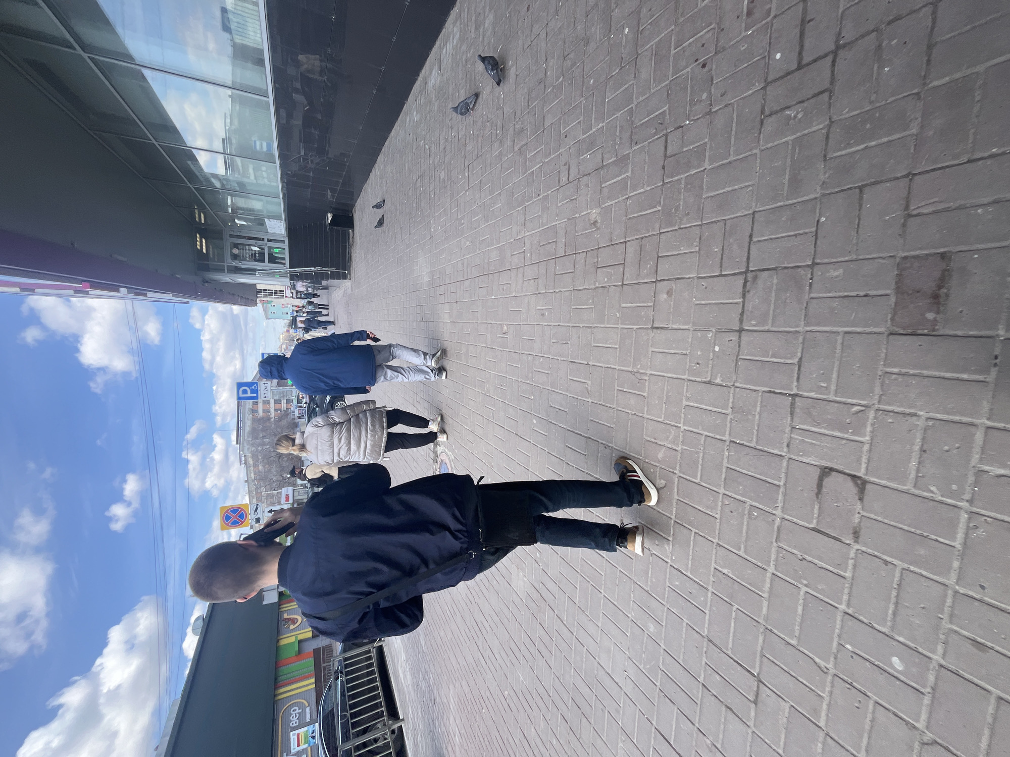 The image depicts a street scene in Ulyanovsk, Russia. The ground is paved with grey rectangular bricks, and a building with reflective windows borders the left side of the frame. Several pigeons are visible on the pavement near the building. Three people are walking in the same direction away from the camera. The person closest to the camera is a man with short hair wearing a navy jacket, black pants, and sneakers with tan accents. He is holding a phone to his ear. Behind him is a person wearing a beige jacket and another person wearing a blue jacket and grey pants. The sky is blue with scattered white clouds. A street sign and a building are visible in the distance. A metal railing and a glimpse of a car are seen along the bottom left edge of the image.
