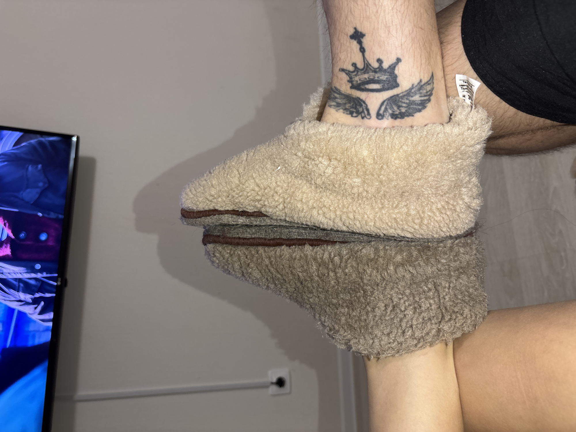 A close-up shot shows a person wearing a tan, plush slipper on one foot. The person has a tattoo of a crown with wings on their ankle. Part of the person's leg is visible, with a black article of clothing near the top of the frame. A reflection of the slipper is seen below. The background includes a television screen to the left and a light-colored wall. There is an electrical outlet on the wall. There is no indication that the image was taken in Izhevsk, Russia.