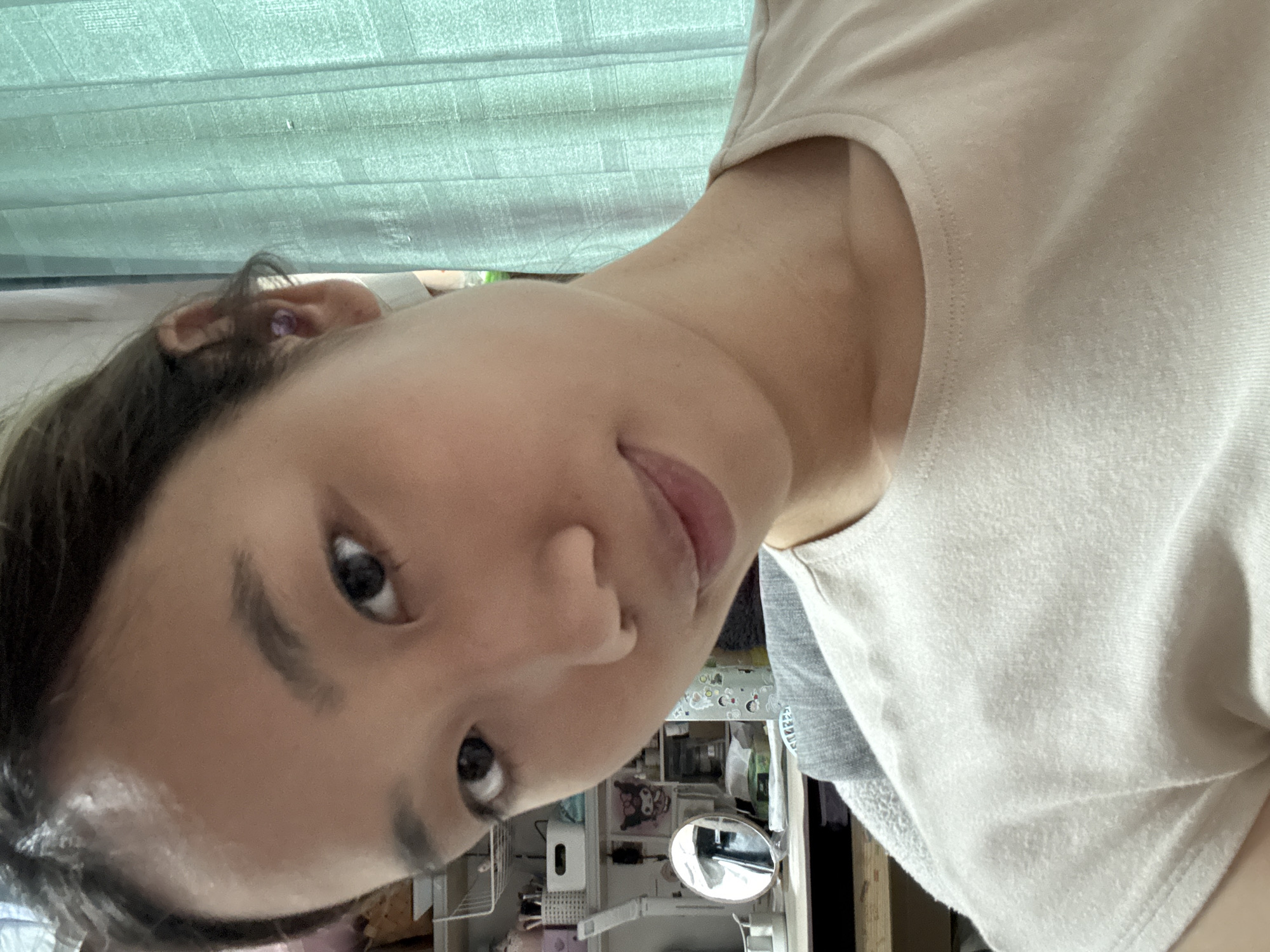 A person is depicted indoors. The person is wearing a light-colored t-shirt and has short, dark hair. They appear to be in a room with a pale green curtain in the background. Various objects are visible on a shelf or surface behind them, including what appears to be a small mirror. The location is possibly within Nan'an District, China.