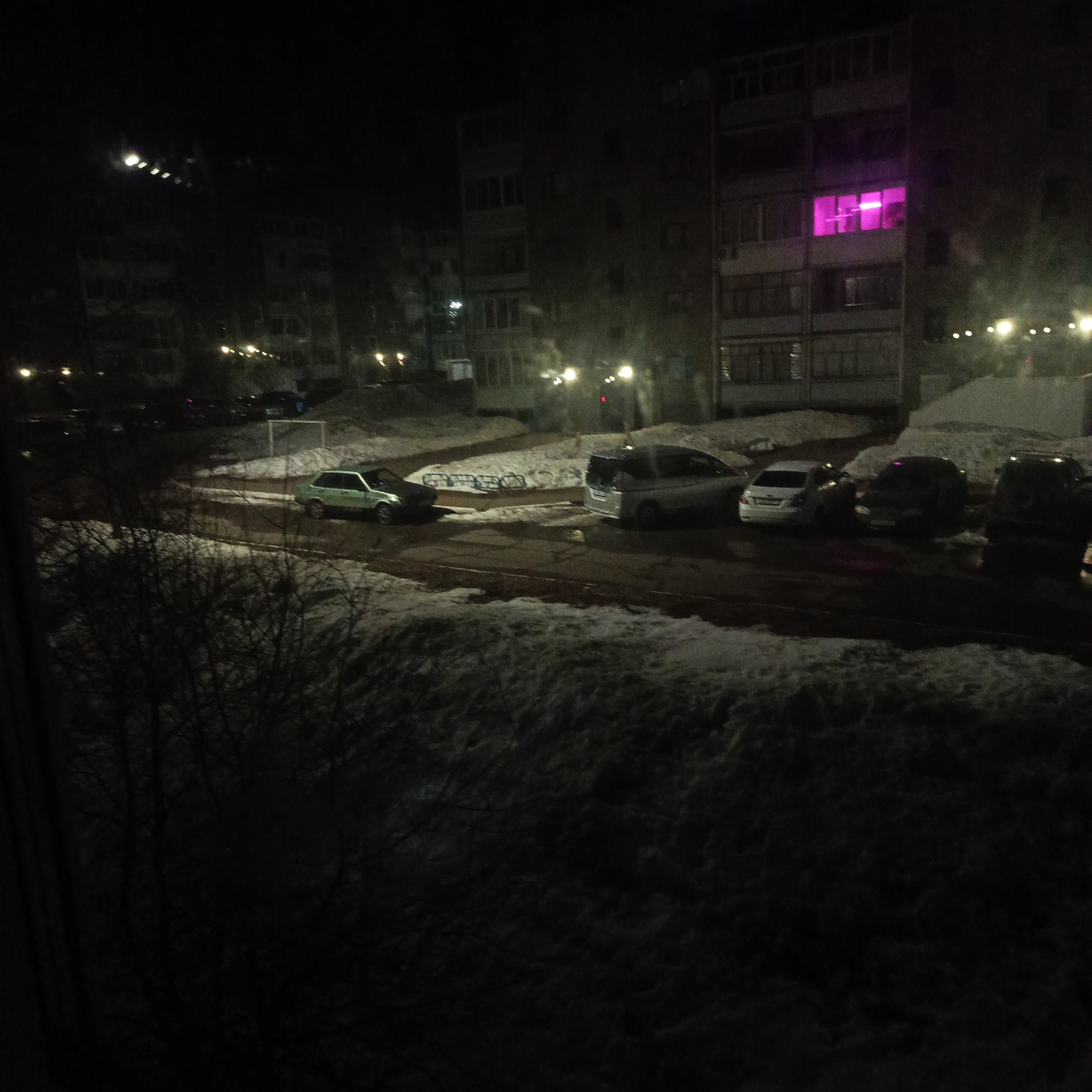 Captured at night, the scene depicts an urban setting in Марковский, Russia. The view is from a window, with a focus on residential buildings and a parking area. The buildings are multi-storied, with one apartment showing a distinct pink glow from its windows. Several cars are parked along the road and in the parking lot, with snow lining the ground and rooftops. Streetlights provide illumination, casting shadows and highlighting the stark contrast between the darkness and light. Bare tree branches are visible in the foreground, adding to the wintry atmosphere. A soccer field is partially visible in the background.