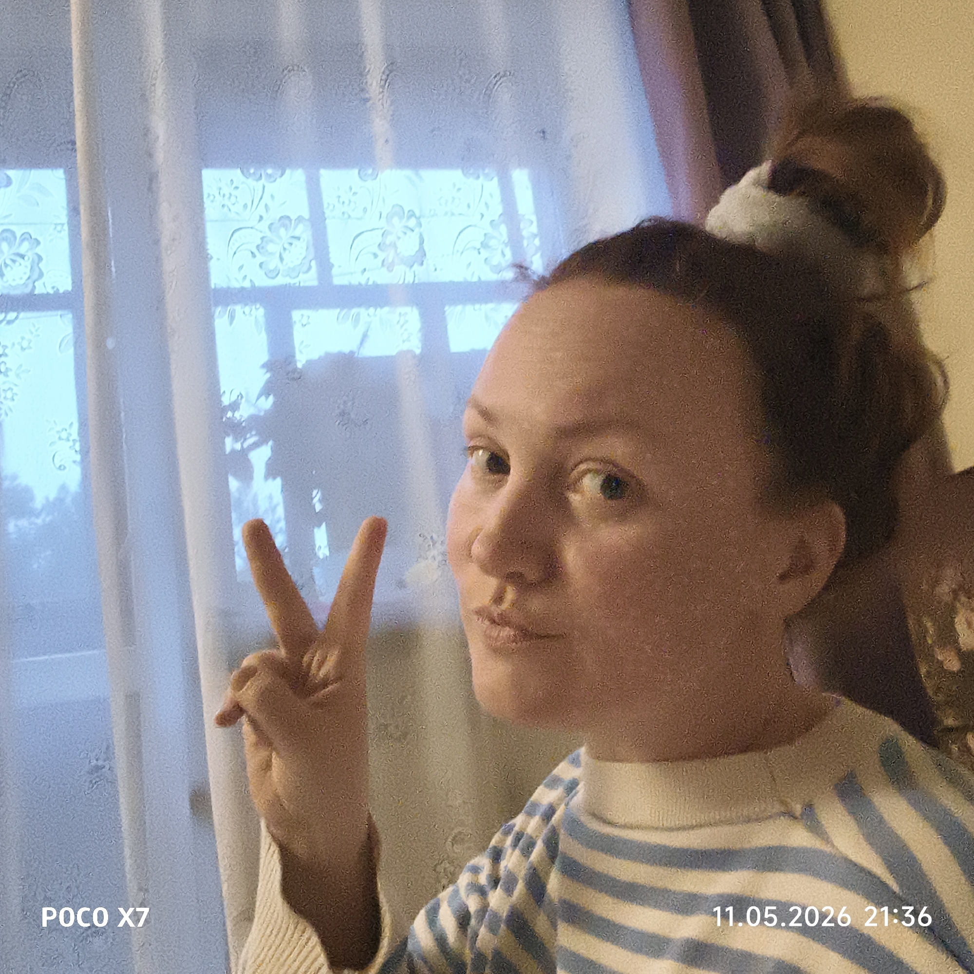 A woman with light skin, light brown hair in a bun, and wearing a blue and white striped shirt is holding up a peace sign with her left hand. The woman is positioned indoors with a large window in the background. The window is covered with a translucent curtain which has flower designs on it. Through the curtain, foliage and a blurred view of the outdoor environment can be seen. A dark purple curtain can be seen slightly to the right of the woman. A stamp is in the lower left of the image that reads "POCO X7". A timestamp in the lower right of the image reads "11.05.2026 21:36". There is no explicit indication that the location is Chaikovsky, Russia.
